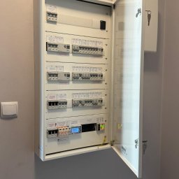 ENERGY - Rozdzielnica elektryczna natynkowa z otwartymi drzwiczkami, widoczne zabezpieczenia, wyłączniki, liczniki. Instalacja w kolorze białym na szarej ścianie.