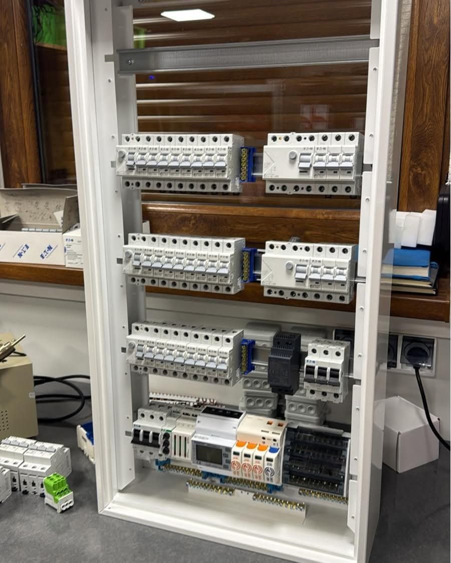 Rozdzielnica elektryczna w białej obudowie, widoczne zabezpieczenia różnicowoprądowe, bezpieczniki i moduły. Elementy instalacji elektrycznej wewnątrz budynku.