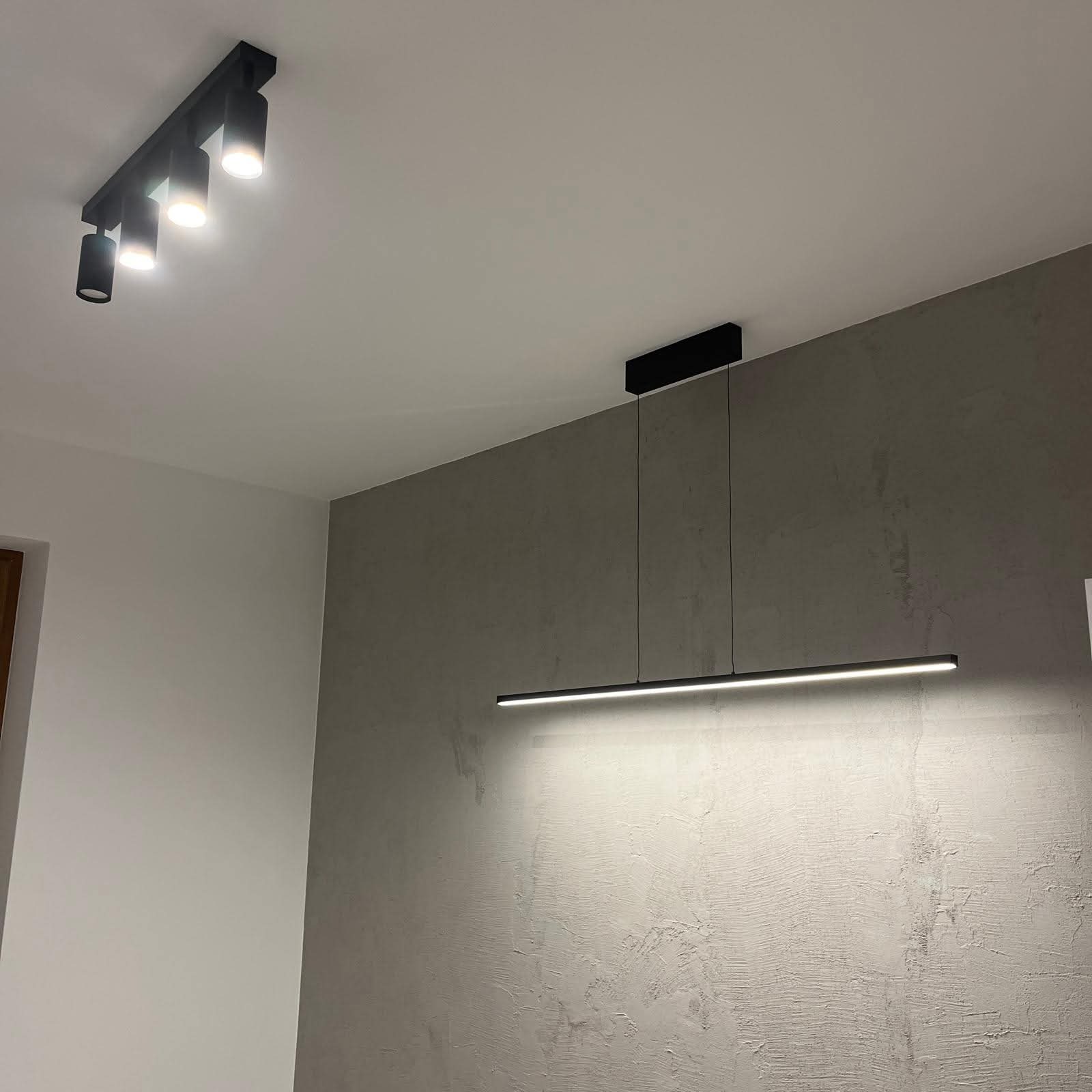Nowoczesne oświetlenie sufitowe: czarna listwa LED zawieszona na tle szarej ściany z fakturą, uzupełniona reflektorami punktowymi. Minimalistyczny design.