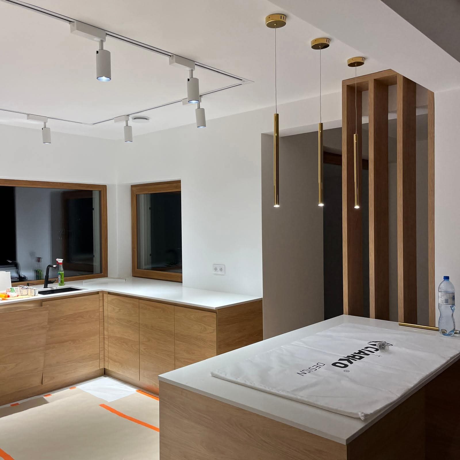 Nowoczesna kuchnia z drewnianymi szafkami, białym blatem i złotymi lampami wiszącymi. Widoczne szyny z reflektorami sufitowymi i dekoracyjna ścianka działowa.