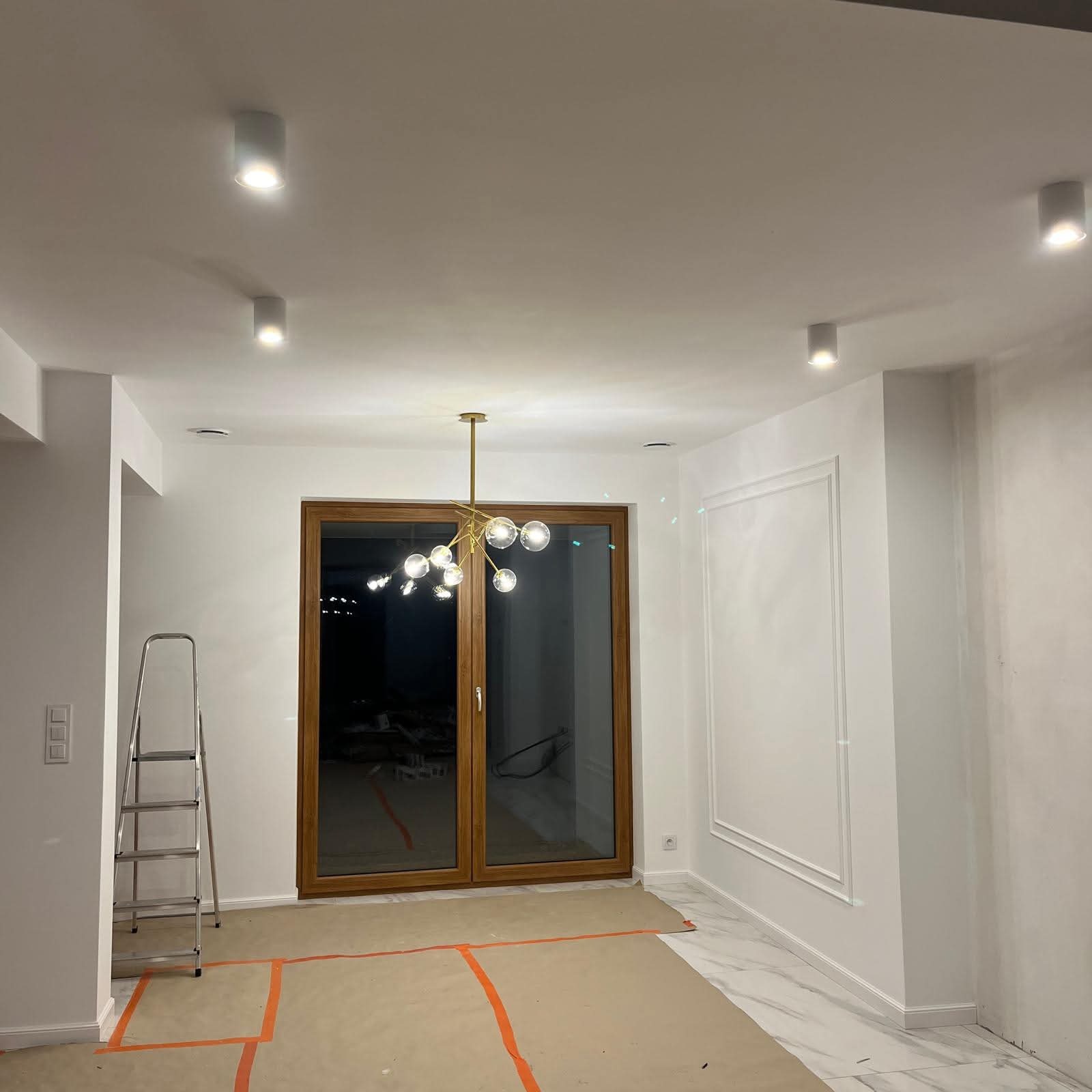 Nowoczesny salon z zamontowanym złotym żyrandolem i białymi lampami sufitowymi. Na podłodze zabezpieczenia malarskie, drabina oparta o ścianę.