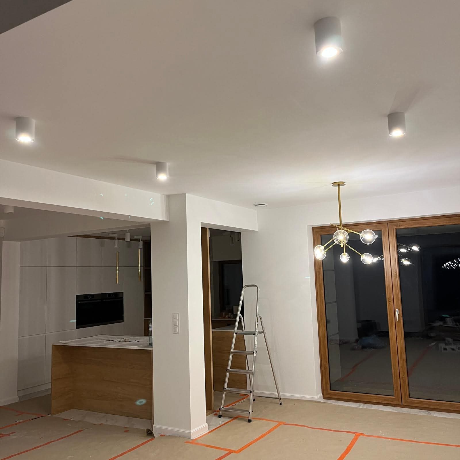 Nowoczesne oświetlenie sufitowe w jasnym wnętrzu z drabiną, prawdopodobnie po montażu. Widoczne lampy punktowe i designerski żyrandol. Podłoga zabezpieczona folią.