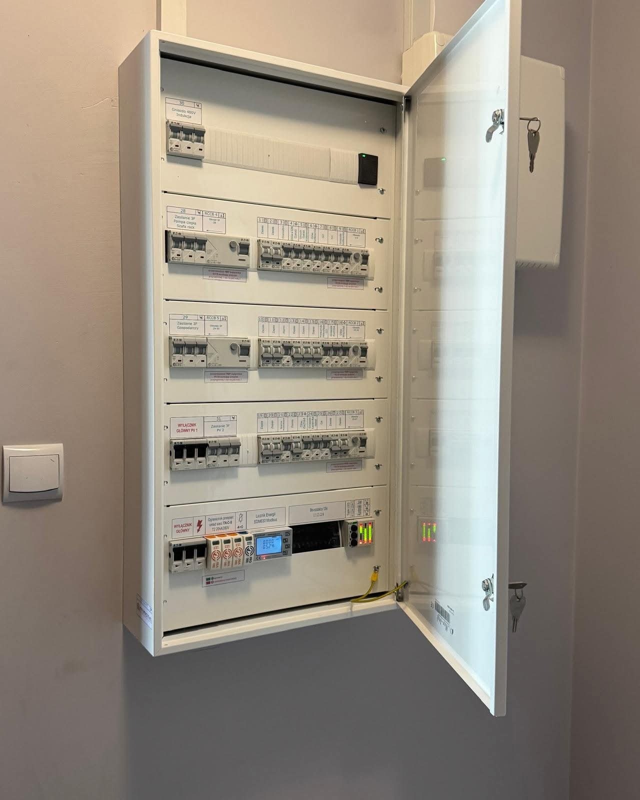 Rozdzielnica elektryczna natynkowa z otwartymi drzwiczkami, widoczne zabezpieczenia, wyłączniki, liczniki. Instalacja w kolorze białym na szarej ścianie.