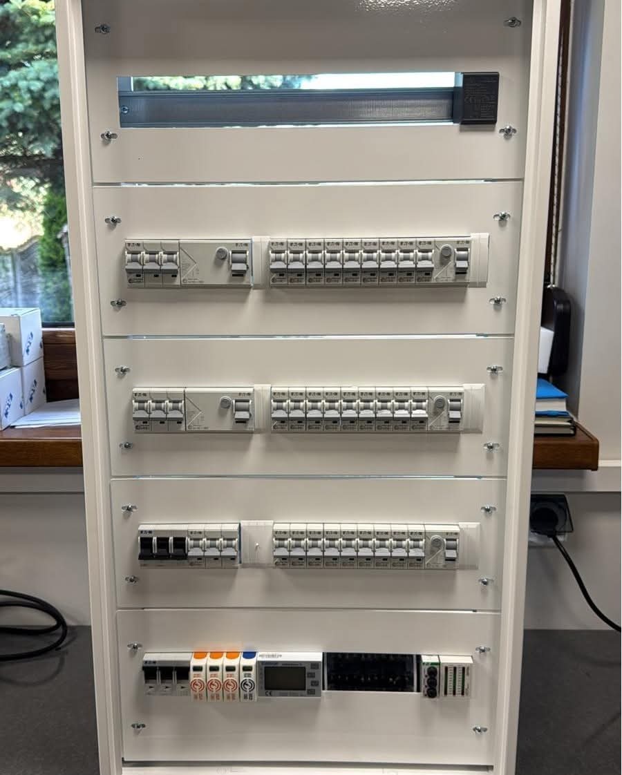 Rozdzielnica elektryczna z zamontowanymi bezpiecznikami i wyłącznikami różnicowoprądowymi, widoczna wnęka na górze, okno w tle.