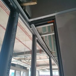 Usługi Transportowe - Montaż aluminiowej ramy okiennej w surowym wnętrzu. Widoczne instalacje pod sufitem i fragmenty płyty karton-gips. Ujęcie z dołu, podkreślające precyzję wykonania.
