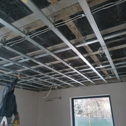 Usługi Transportowe - Montaż metalowej konstrukcji sufitu podwieszanego w pomieszczeniu z oknem. Widoczne profile, kable i częściowo osoba na drabinie. Prace wykończeniowe w trakcie.