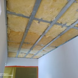 Usługi Transportowe - Sufit podwieszany z izolacją z wełny mineralnej, widok z dołu. Metalowa konstrukcja nośna. Etap prac wykończeniowych w pomieszczeniu z białymi ścianami.