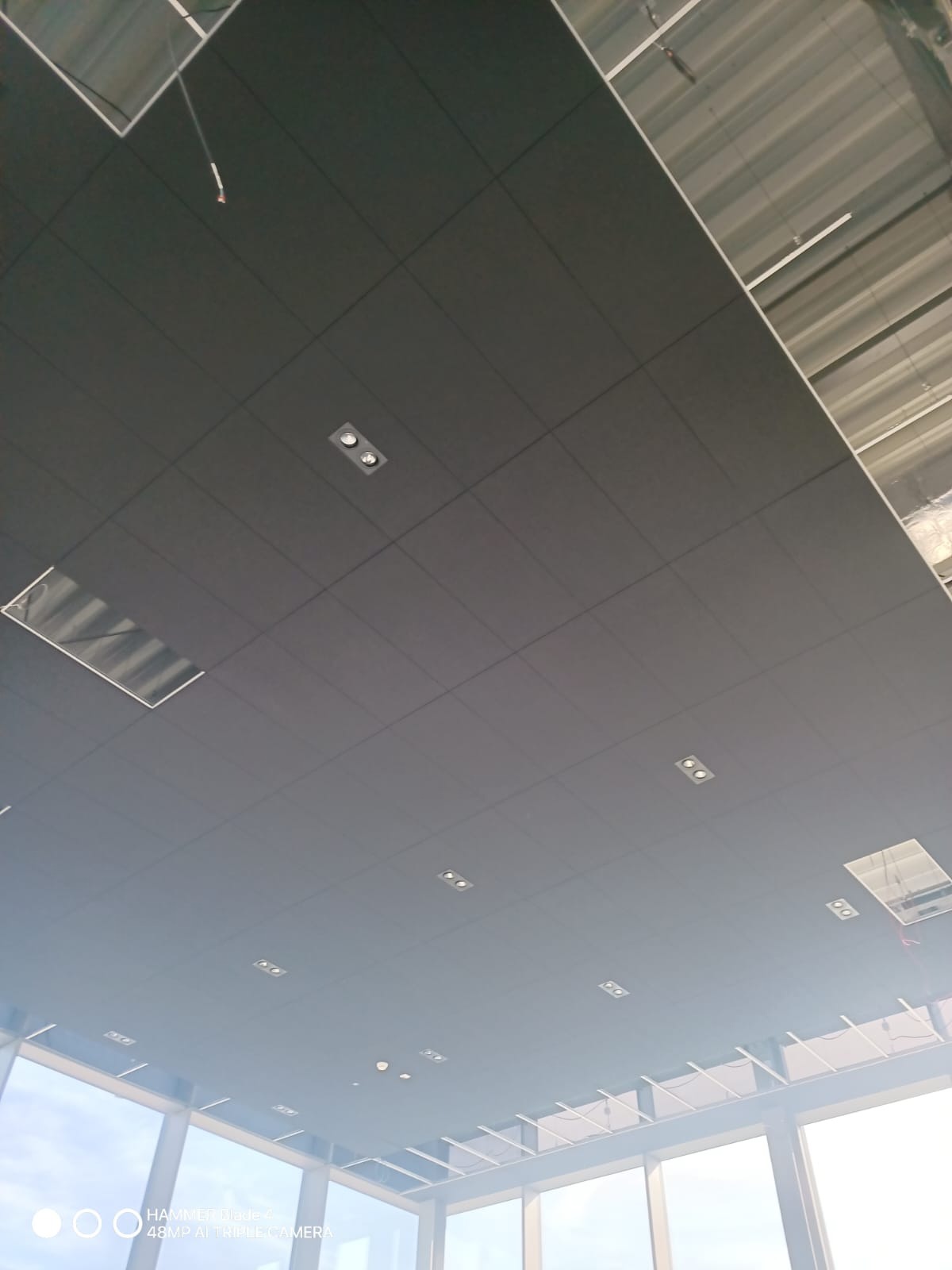 Ciemny sufit podwieszany z wbudowanym oświetleniem punktowym w nowoczesnym biurze z dużymi oknami. Widoczne elementy konstrukcyjne sufitu.