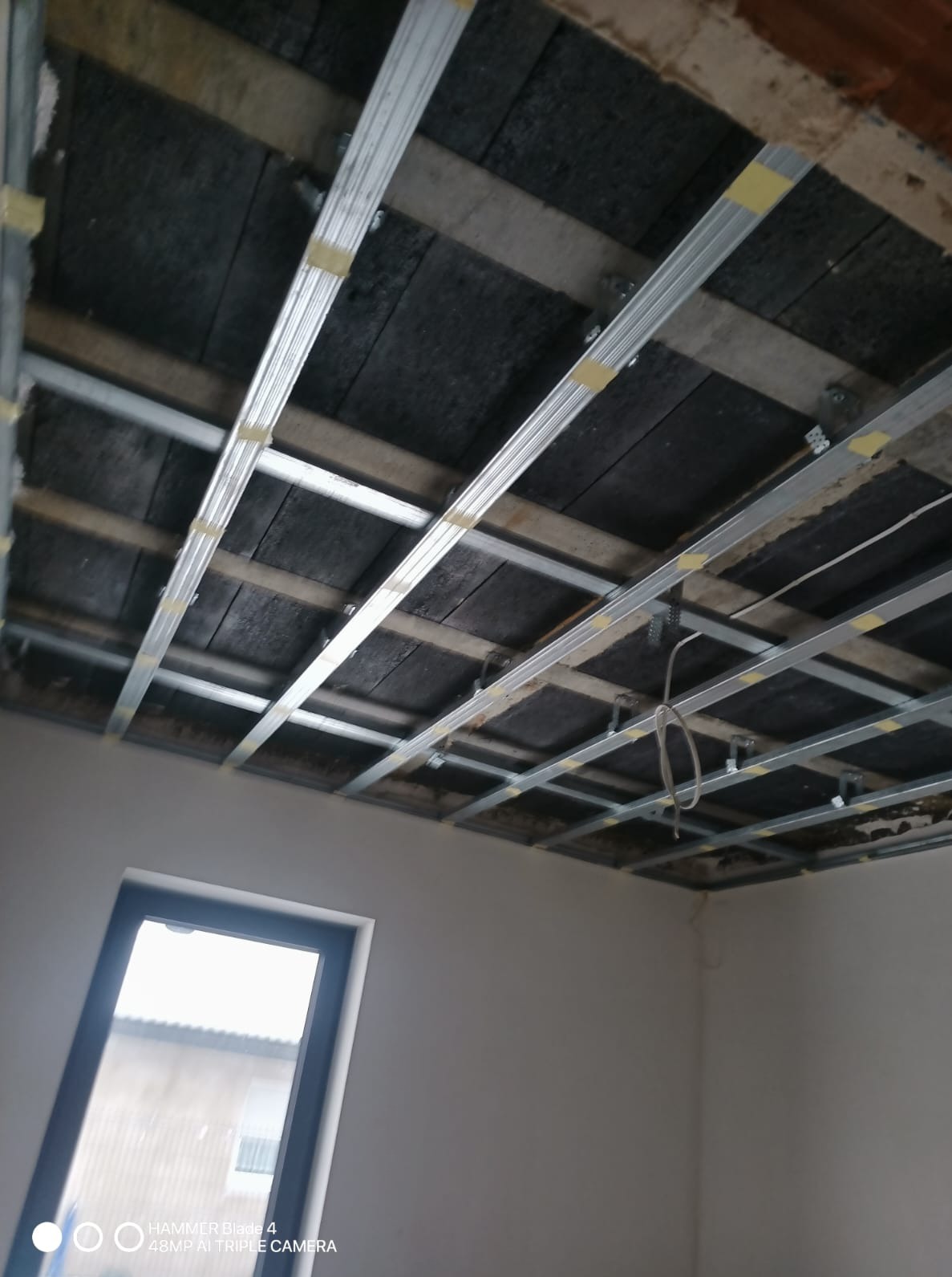 Montaż metalowej konstrukcji sufitu podwieszanego z widocznymi elementami mocującymi i izolacją, w jasnym pomieszczeniu z oknem. Etap prac wykończeniowych.