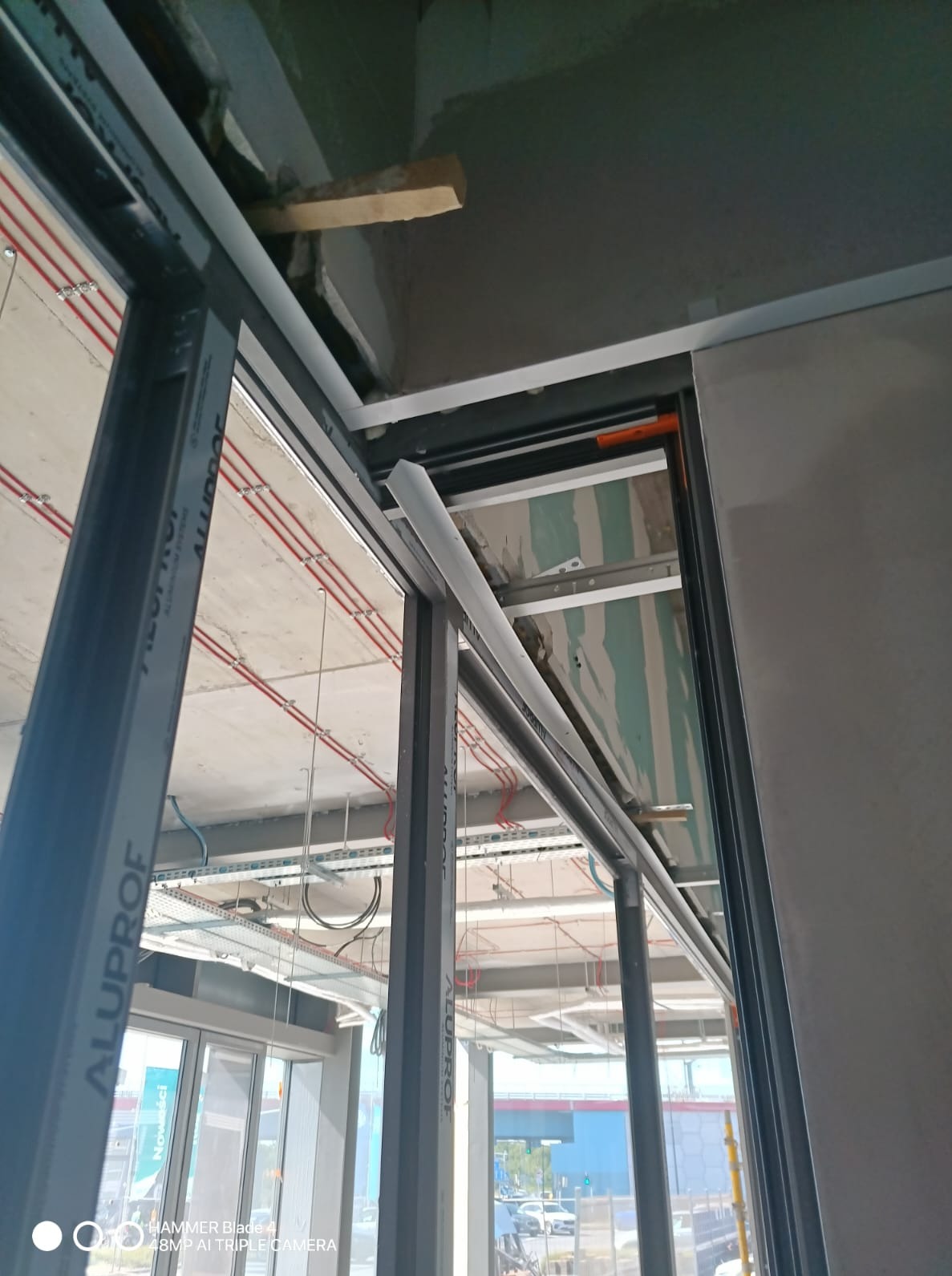 Montaż aluminiowej ramy okiennej w surowym wnętrzu. Widoczne instalacje pod sufitem i fragmenty płyty karton-gips. Ujęcie z dołu, podkreślające precyzję wykonania.