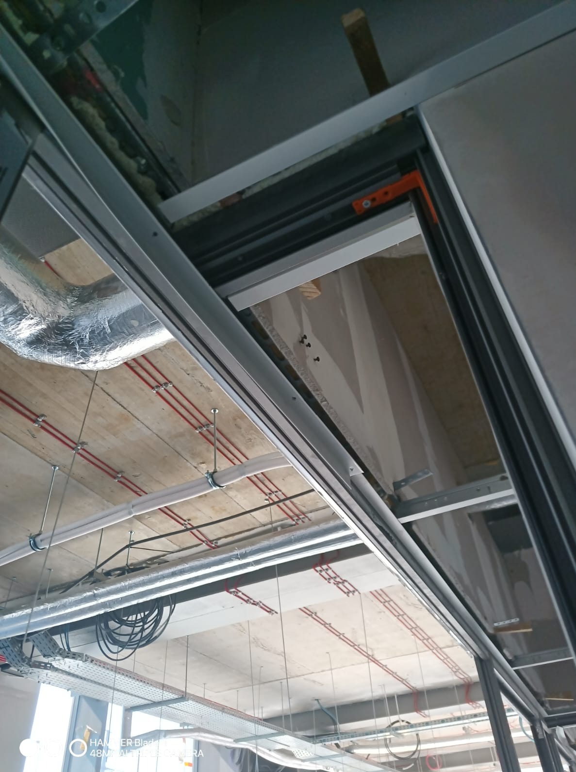 Sufit w trakcie montażu podwieszanego, widoczne instalacje elektryczne, rury wentylacyjne i profile konstrukcyjne. Surowy stan wykończenia, detale mocowań.