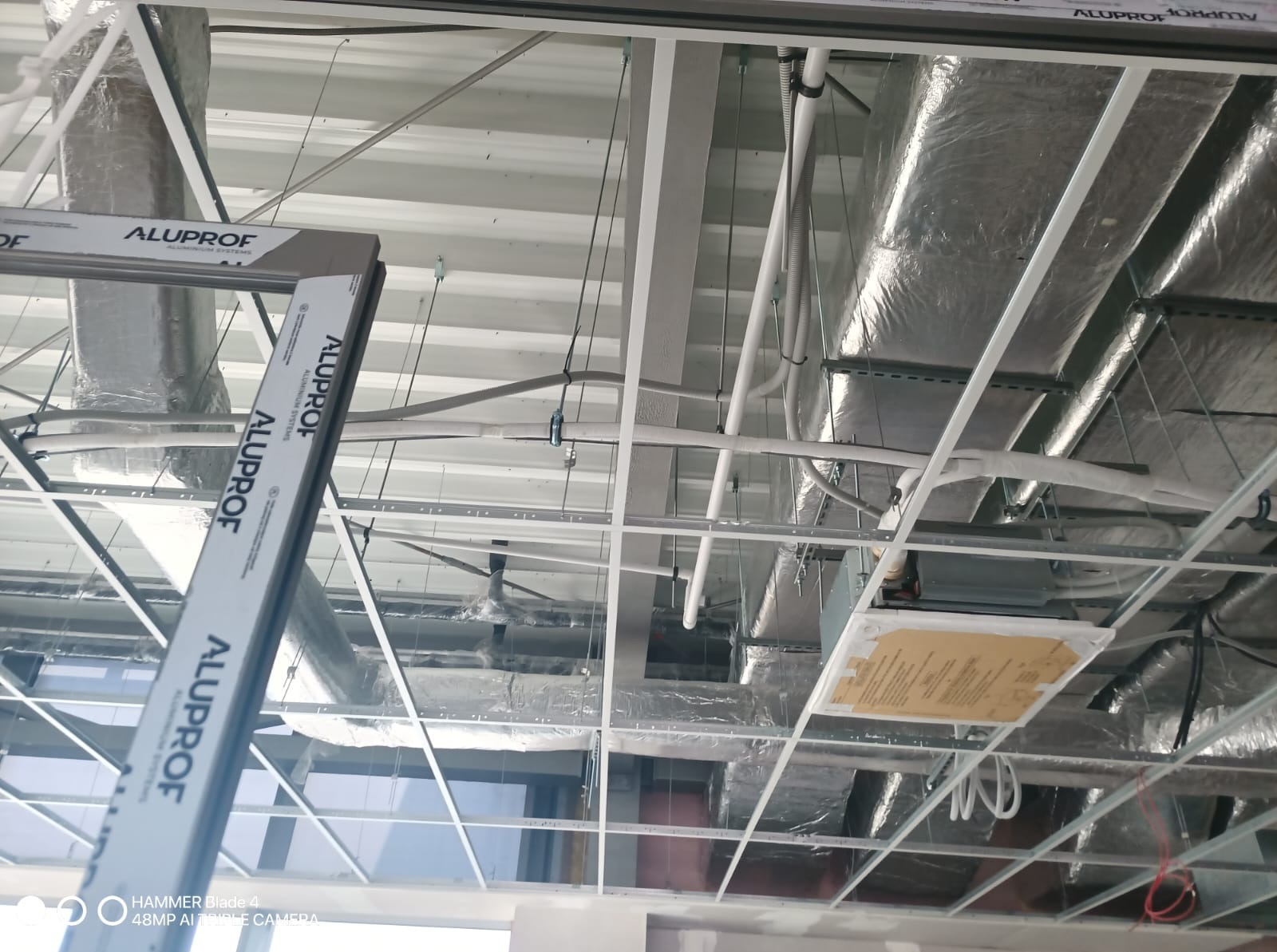 Sufit podwieszany z widoczną konstrukcją metalową, instalacją wentylacyjną i przewodami. Widoczne profile Aluporf. Ujęcie z dołu, perspektywa z wnętrza budynku.