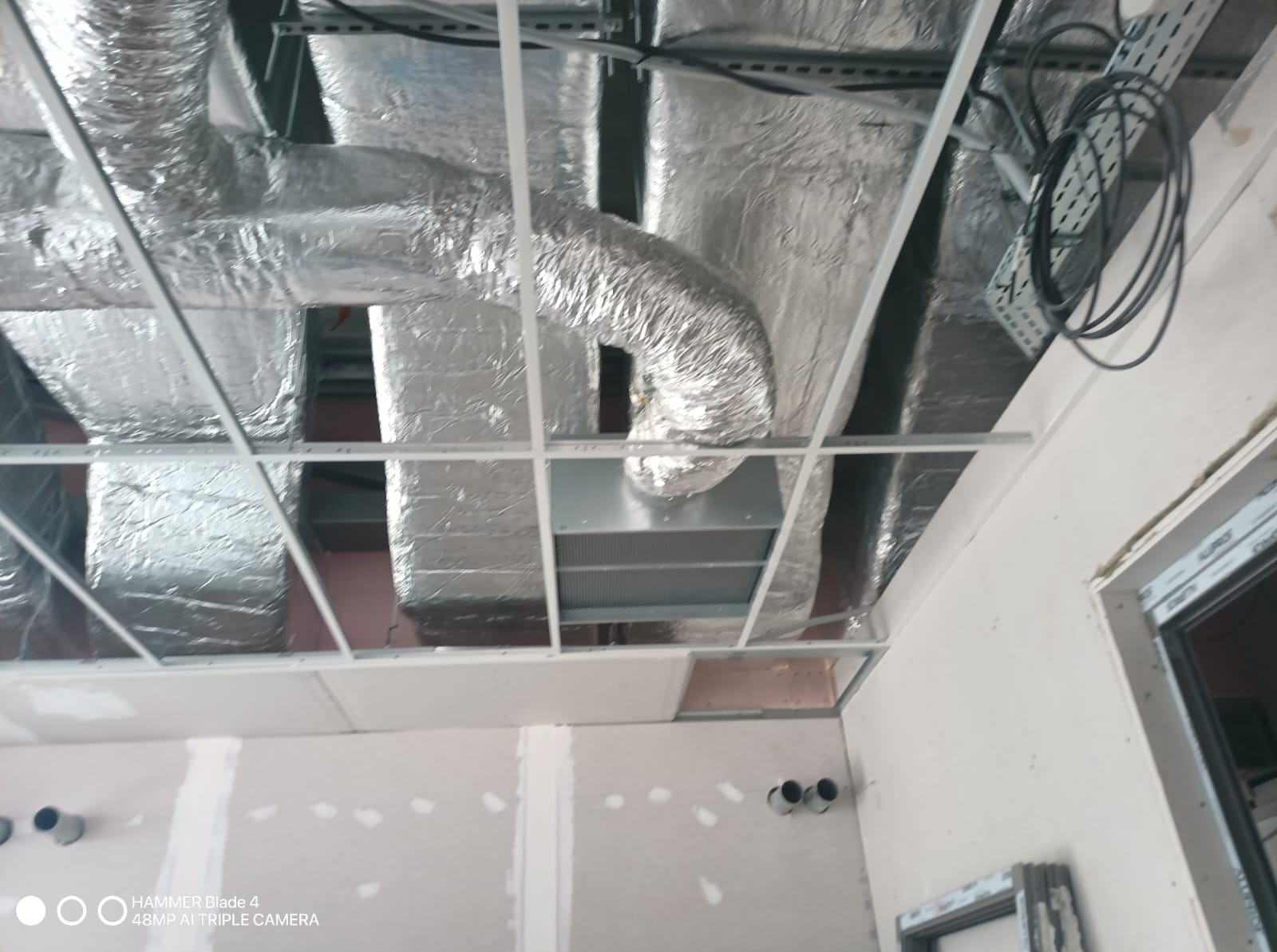 Srebrne rury wentylacyjne pod sufitem podwieszanym w trakcie montażu. Widoczne profile, płyty gipsowe i instalacja elektryczna. Ujęcie z dołu, surowy stan wykończenia.