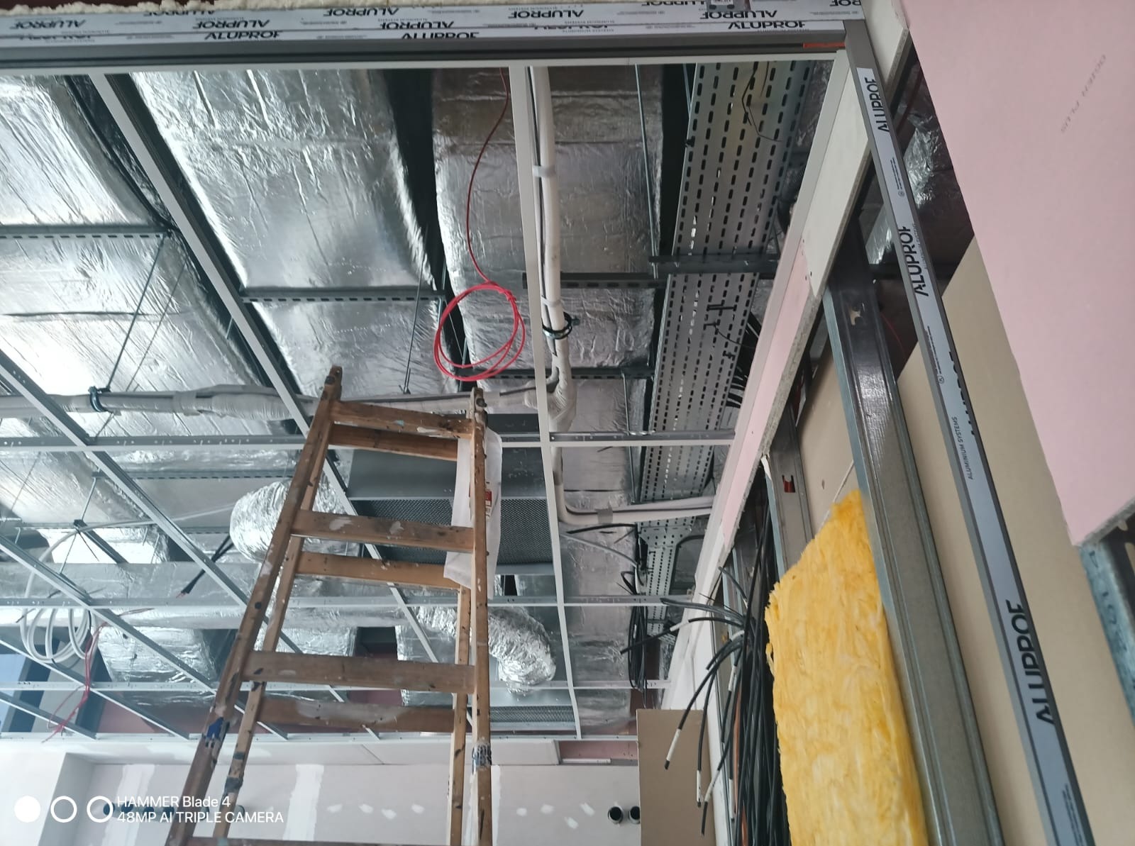 Montaż sufitu podwieszanego z widoczną izolacją, instalacjami i drabina. Prace wykończeniowe w pomieszczeniu z elementami konstrukcji metalowej i płyt kartonowo-gipsowych.