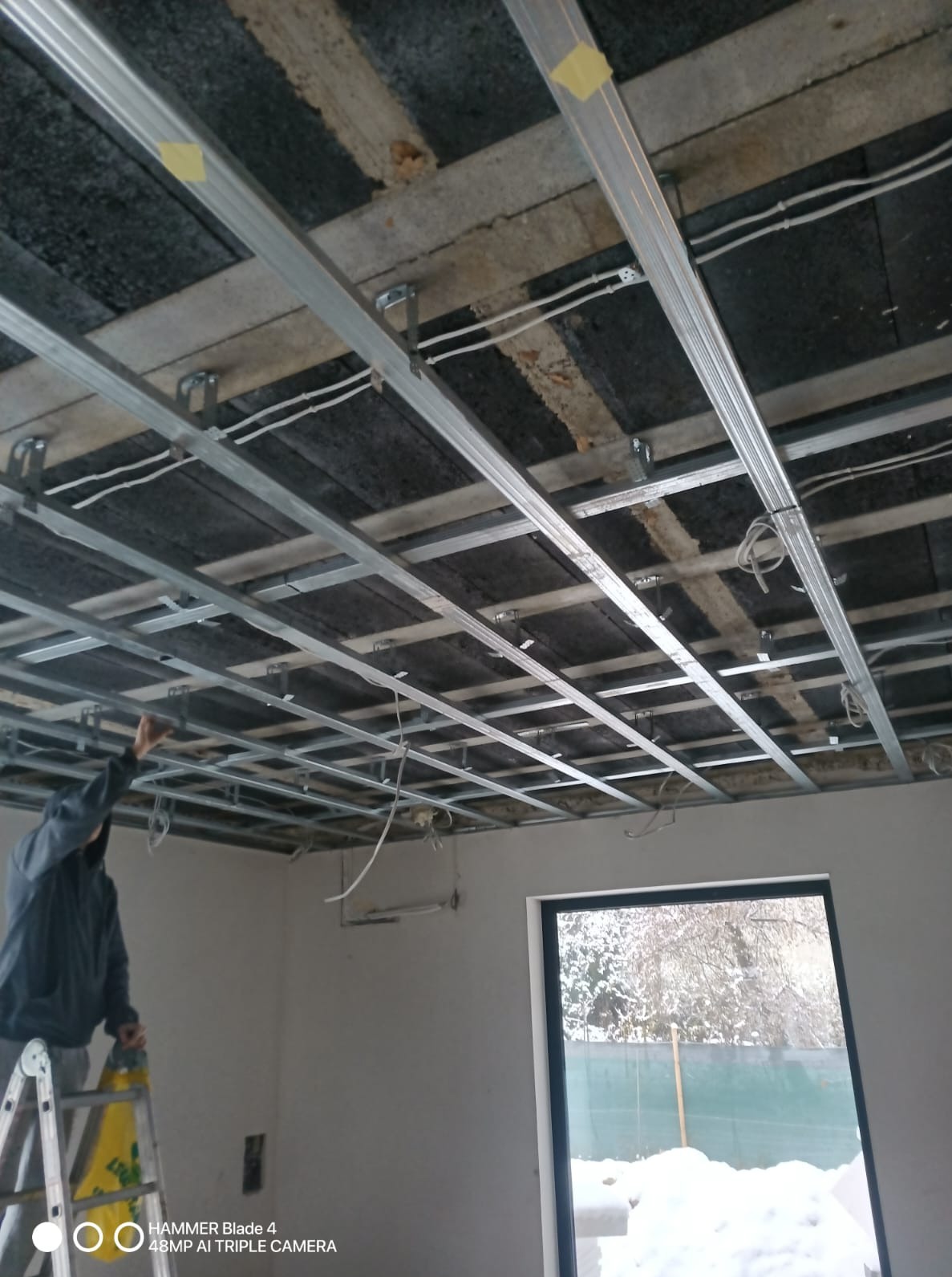 Montaż metalowej konstrukcji sufitu podwieszanego w pomieszczeniu z oknem. Widoczne profile, kable i częściowo osoba na drabinie. Prace wykończeniowe w trakcie.