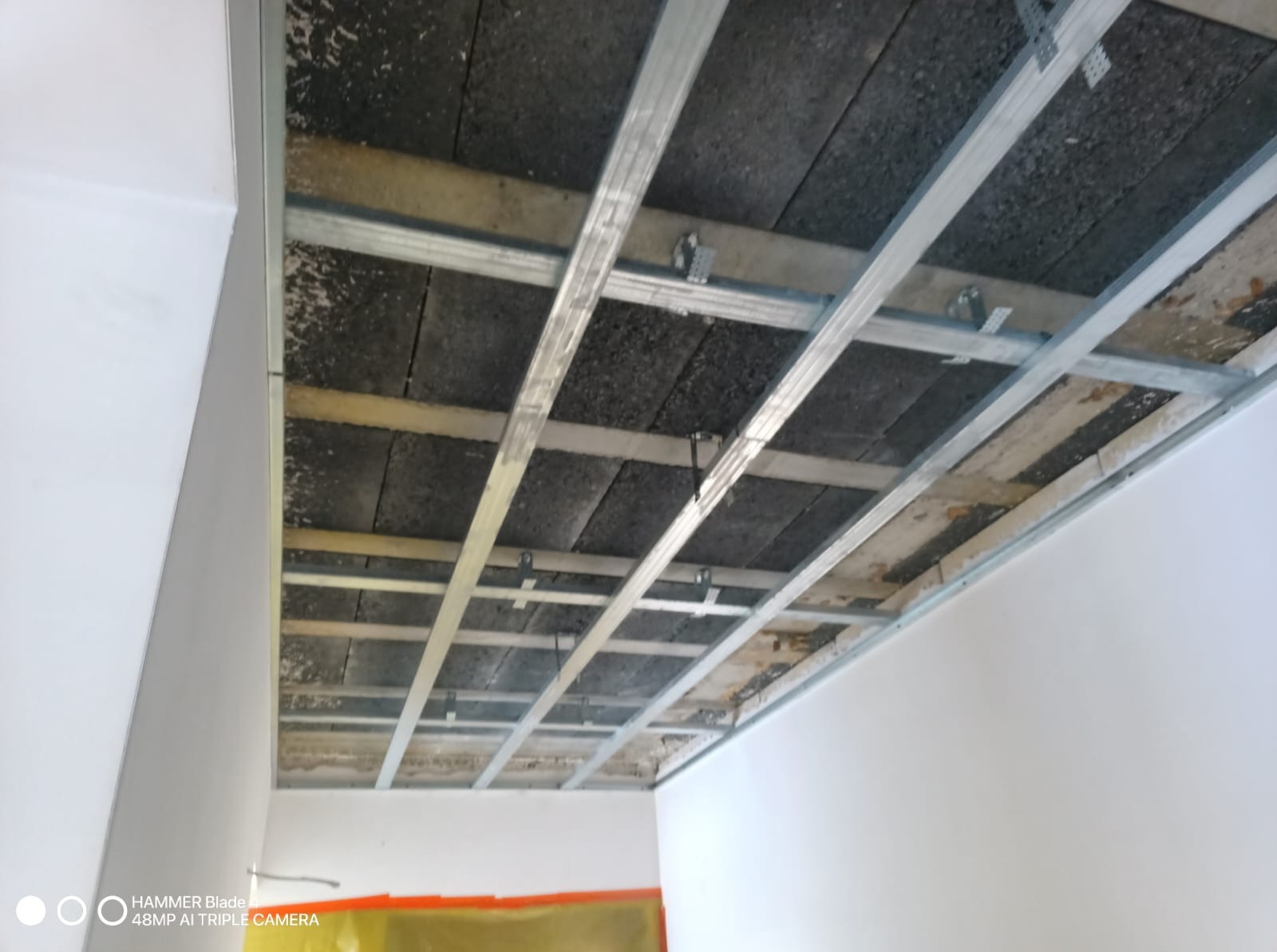 Metalowa konstrukcja sufitu podwieszanego z widoczną izolacją, zamontowana w pomieszczeniu z białymi ścianami. Widok z dołu, sufit w trakcie montażu.