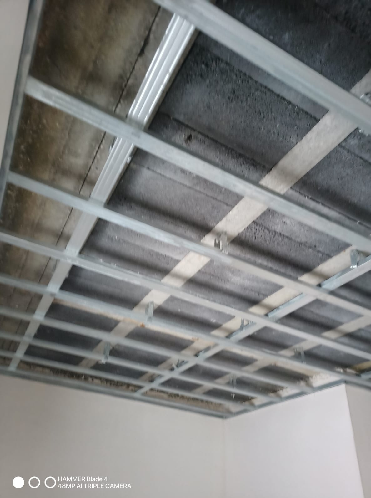 Metalowa konstrukcja sufitu podwieszanego z płyt kartonowo-gipsowych, widok z dołu, surowy stan przed montażem płyt. Szara betonowa powierzchnia w tle.