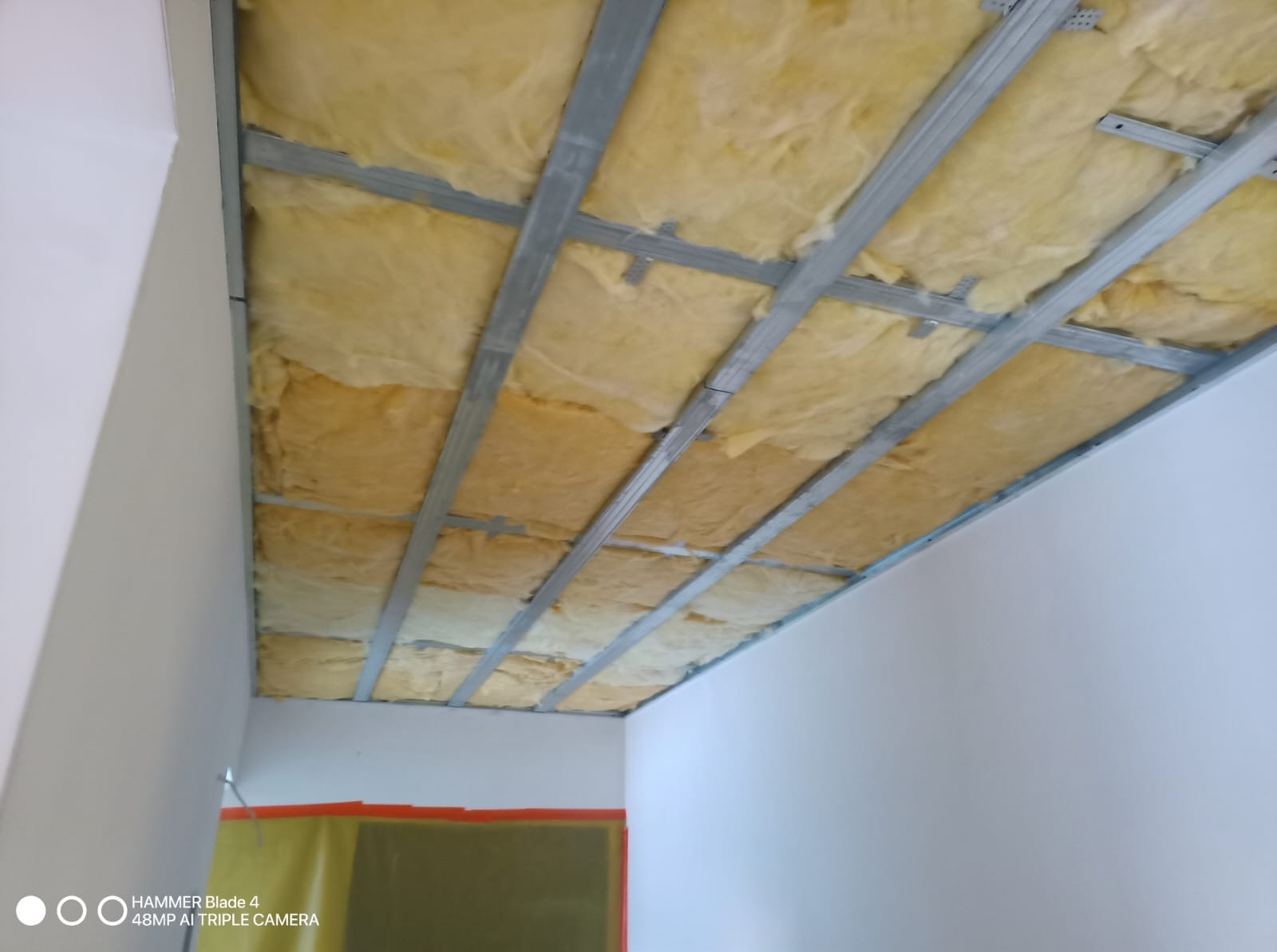 Sufit podwieszany z izolacją z wełny mineralnej, widok z dołu. Metalowa konstrukcja nośna. Etap prac wykończeniowych w pomieszczeniu z białymi ścianami.