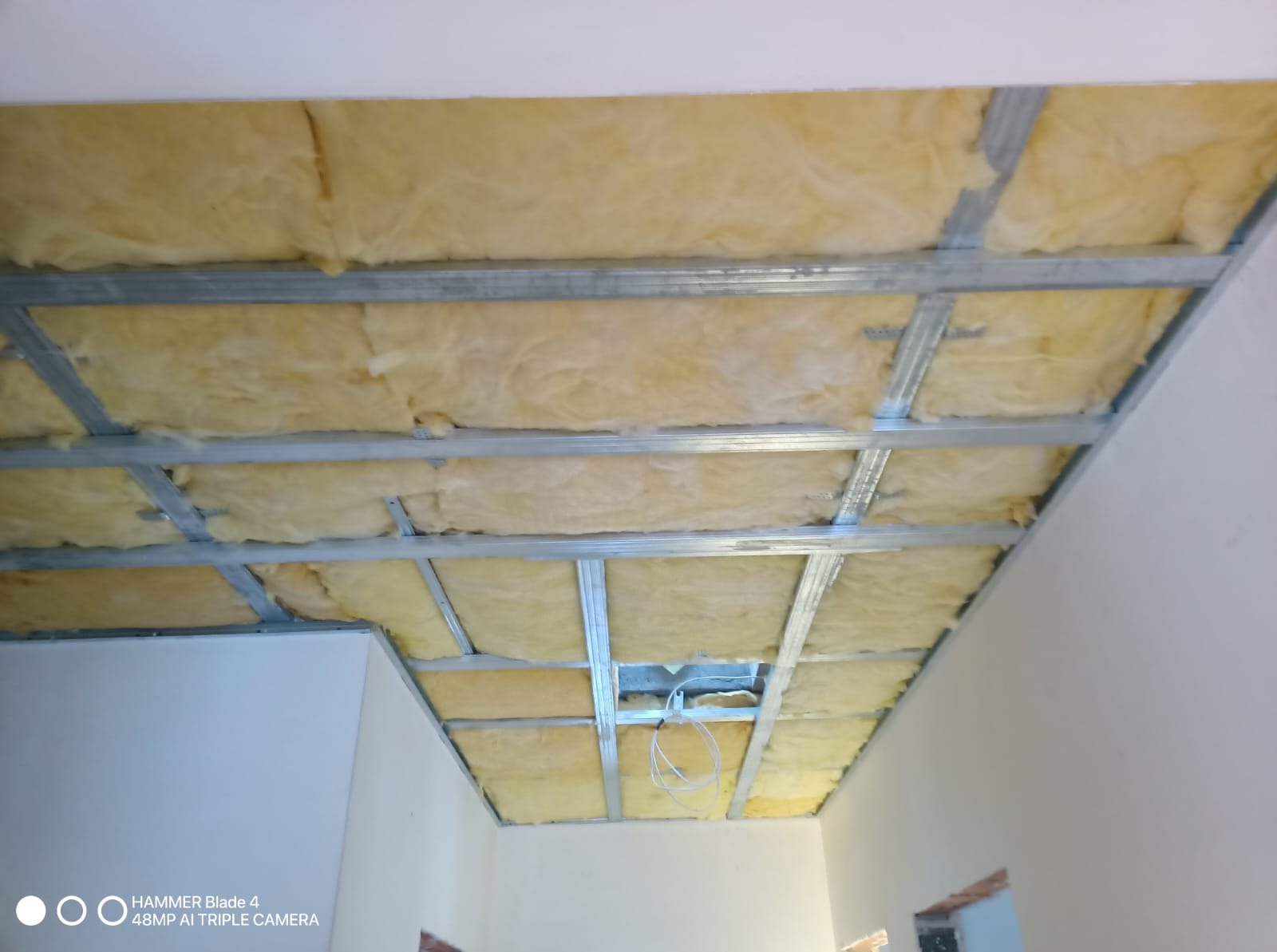 Sufit podwieszany z izolacją z wełny mineralnej, widoczne metalowe profile. Etap prac wykończeniowych, oświetlenie tymczasowe.