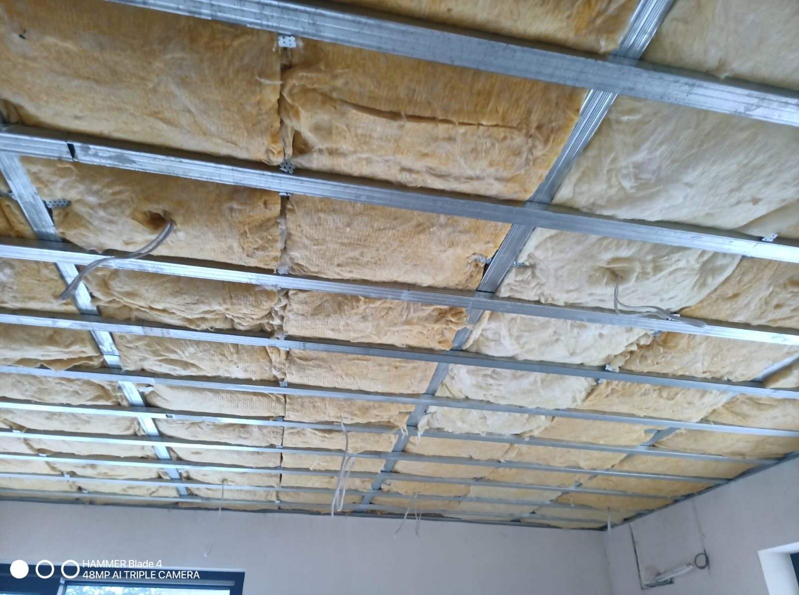 Montaż sufitu podwieszanego z izolacją z wełny mineralnej. Widoczne metalowe profile i instalacja elektryczna. Prace wykończeniowe w nowym budownictwie.