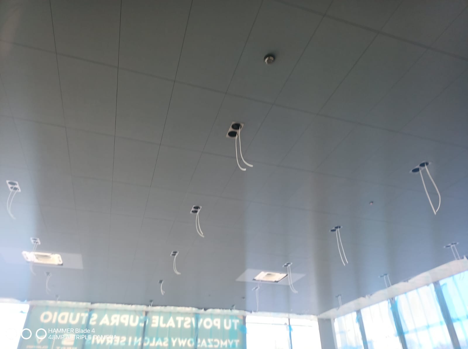 Sufit podwieszany z otworami na oświetlenie w trakcie montażu, widoczne przewody. Prace wykończeniowe w Katowicach. Szara kolorystyka, regularny układ paneli.