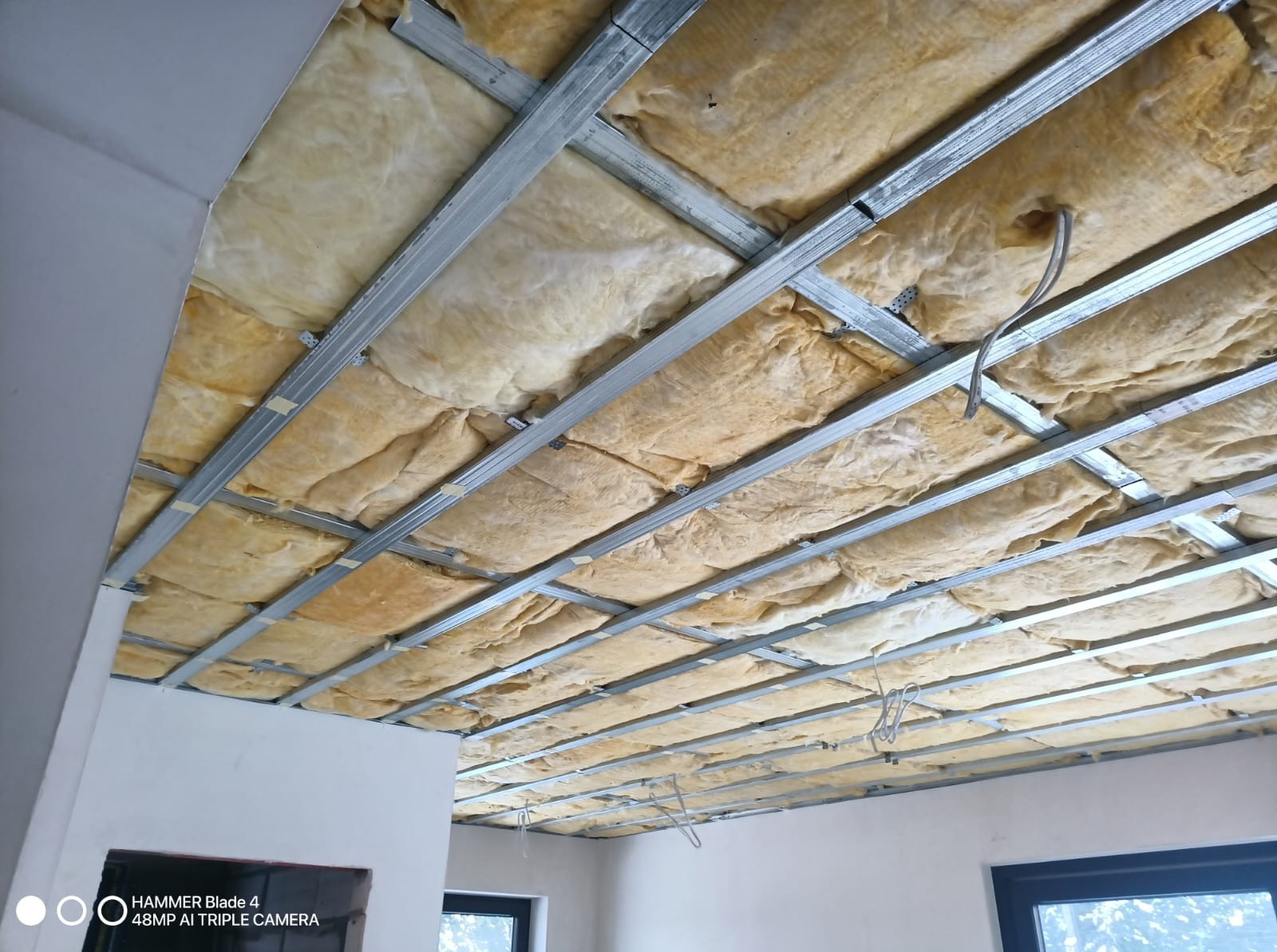 Sufit podwieszany z izolacją z wełny mineralnej, widoczne metalowe profile konstrukcyjne. Etap prac wykończeniowych, instalacja oświetlenia w tle.