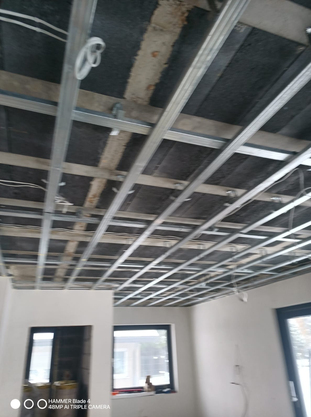 Sufit podwieszany z metalową konstrukcją w trakcie montażu, widoczne przewody elektryczne. Surowy stan wykończenia wnętrza, okna z widokiem na zewnątrz.