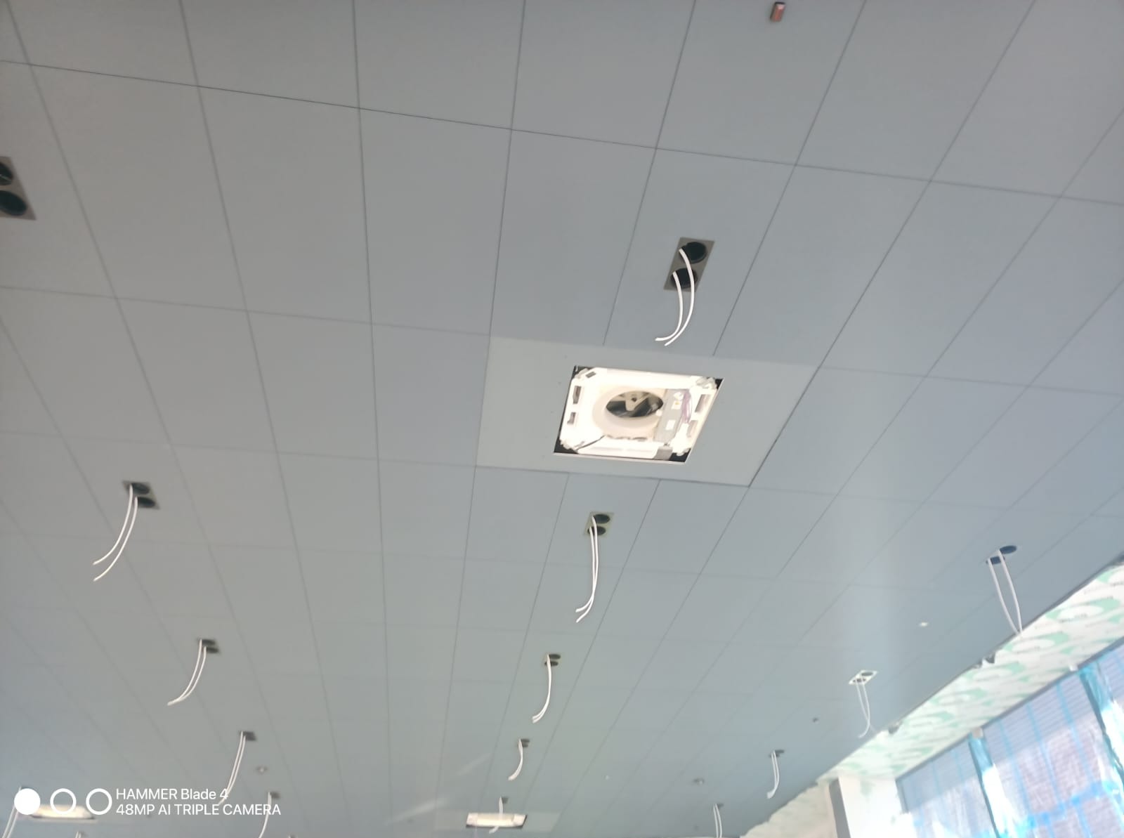 Sufit podwieszany kasetonowy z otworami na oświetlenie i wentylację, widok z dołu, przewody elektryczne wystające z otworów, jasna kolorystyka.