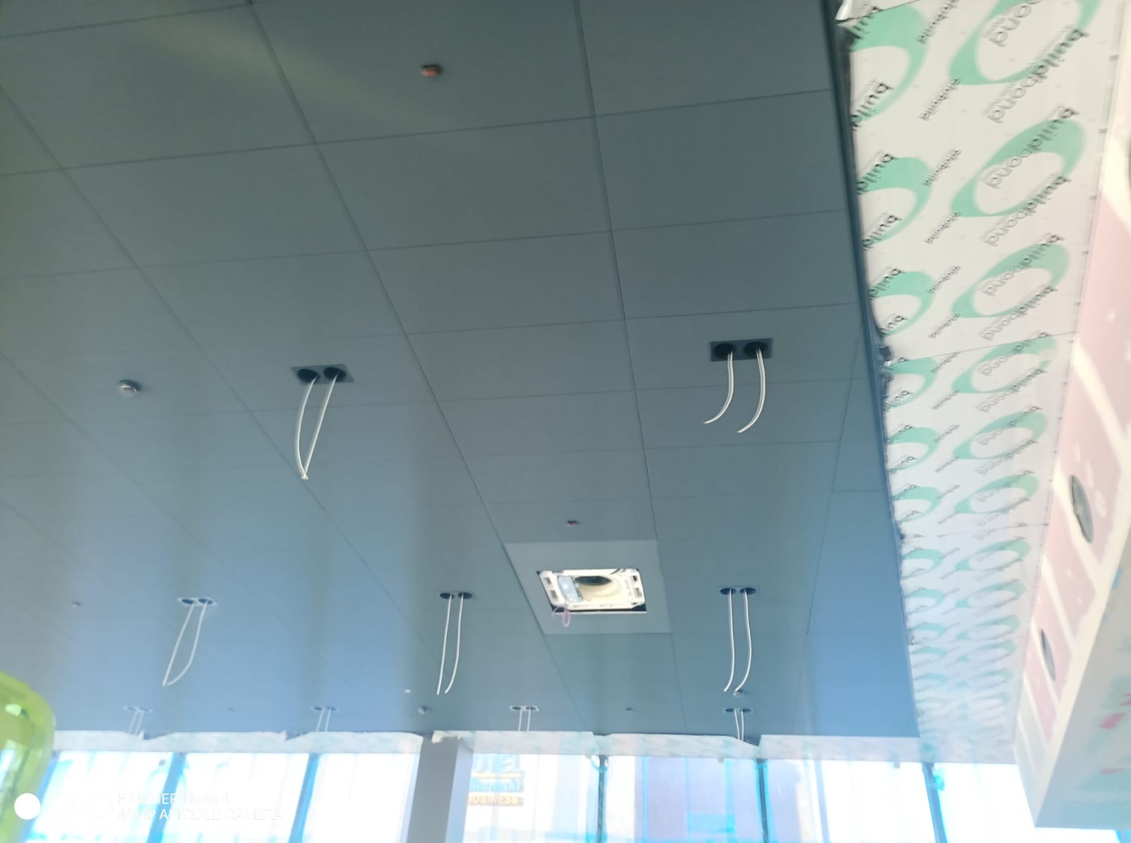 Sufit podwieszany z płyt z widocznymi przewodami elektrycznymi i otworem na klimatyzację. Widok z dołu, w tle okna.