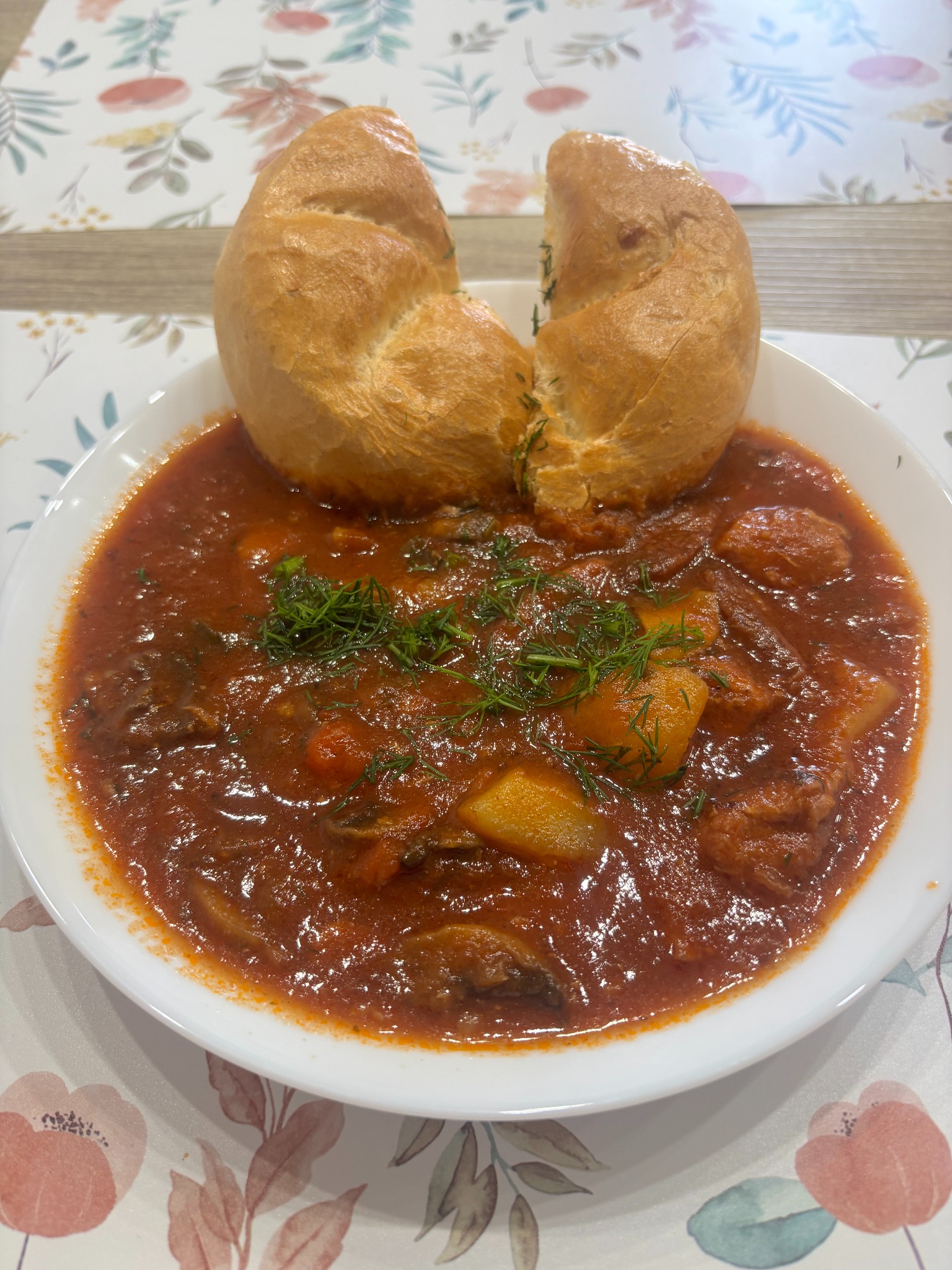 Aromatyczny gulasz z wołowiny i warzyw w białej misce, udekorowany świeżym koperkiem, podany z pszenną bułką na kwiecistym obrusie.