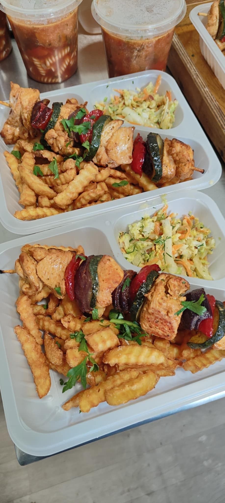 Dwa zestawy cateringowe w plastikowych pojemnikach: szaszłyki z kurczaka i warzyw, frytki karbowane i surówka. Dodatkowo zupy w plastikowych pojemnikach.