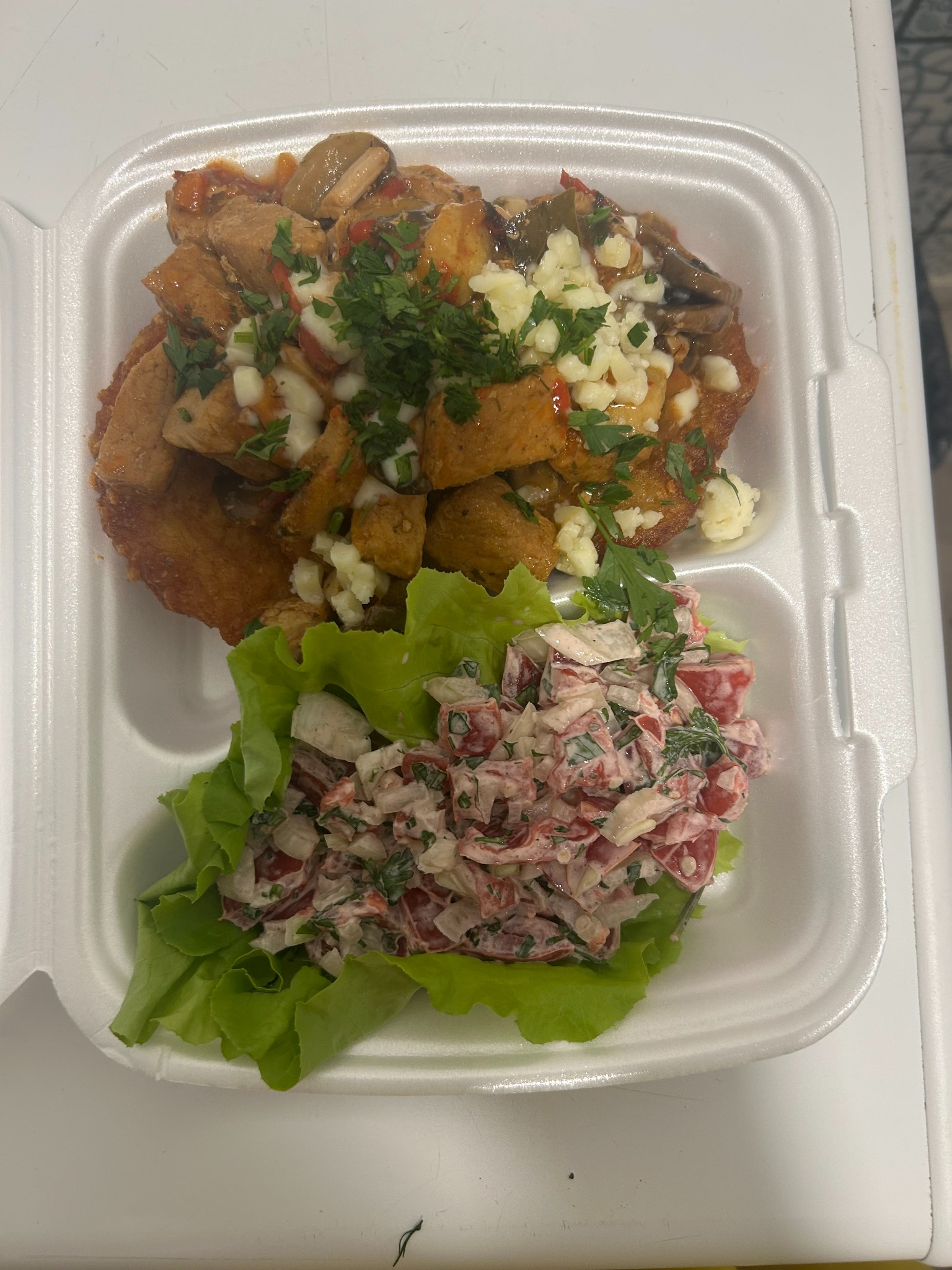 Danie obiadowe w białym pojemniku: kotlet z sosem, posypany serem i natką pietruszki, obok sałatka warzywna na liściach sałaty. Gotowe danie na wynos.