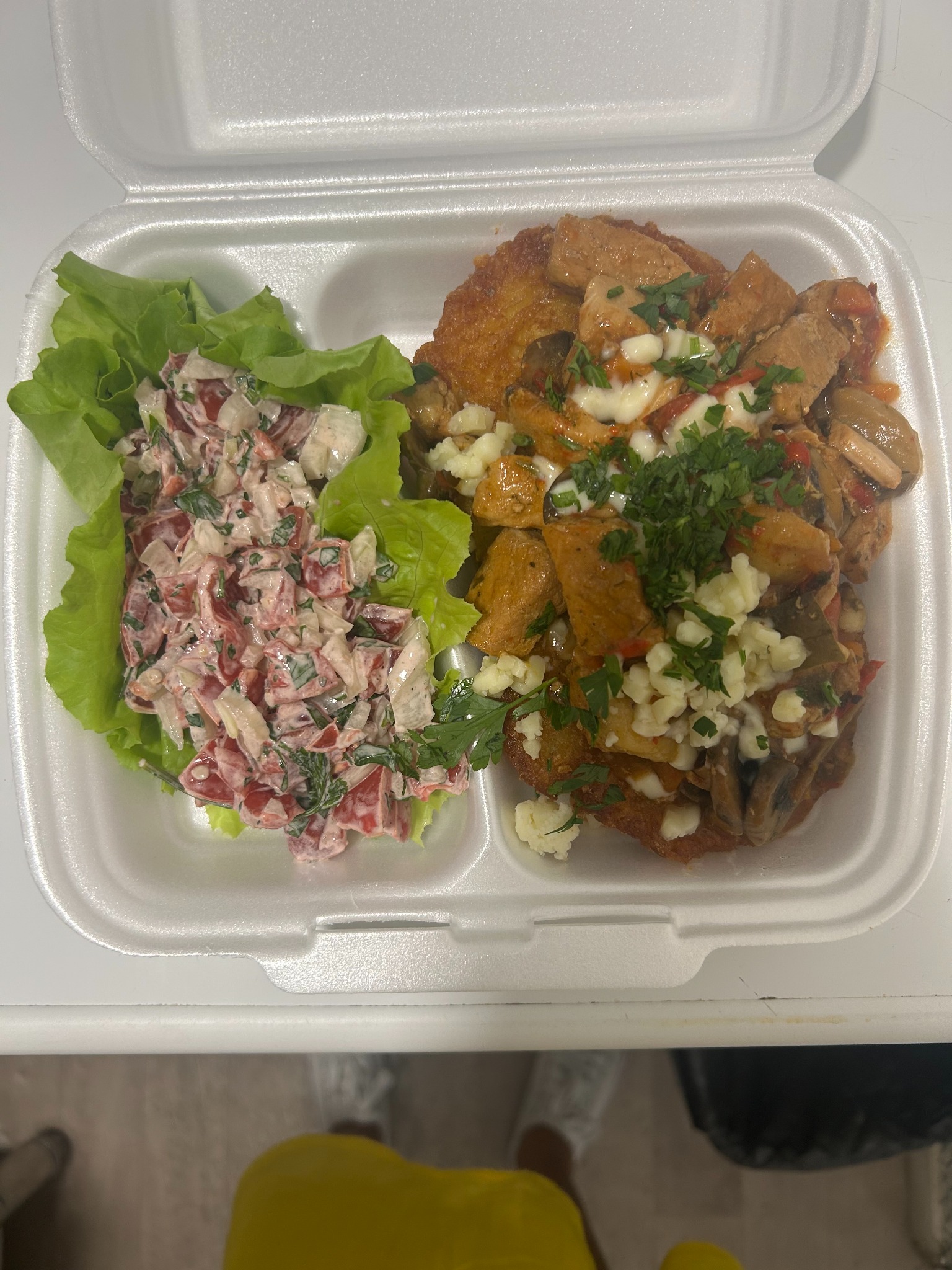 Danie obiadowe w białym pojemniku styropianowym: kotlet z sosem mięsnym, grzybami i serem, obok sałatka warzywna na liściach sałaty. Widok z góry.