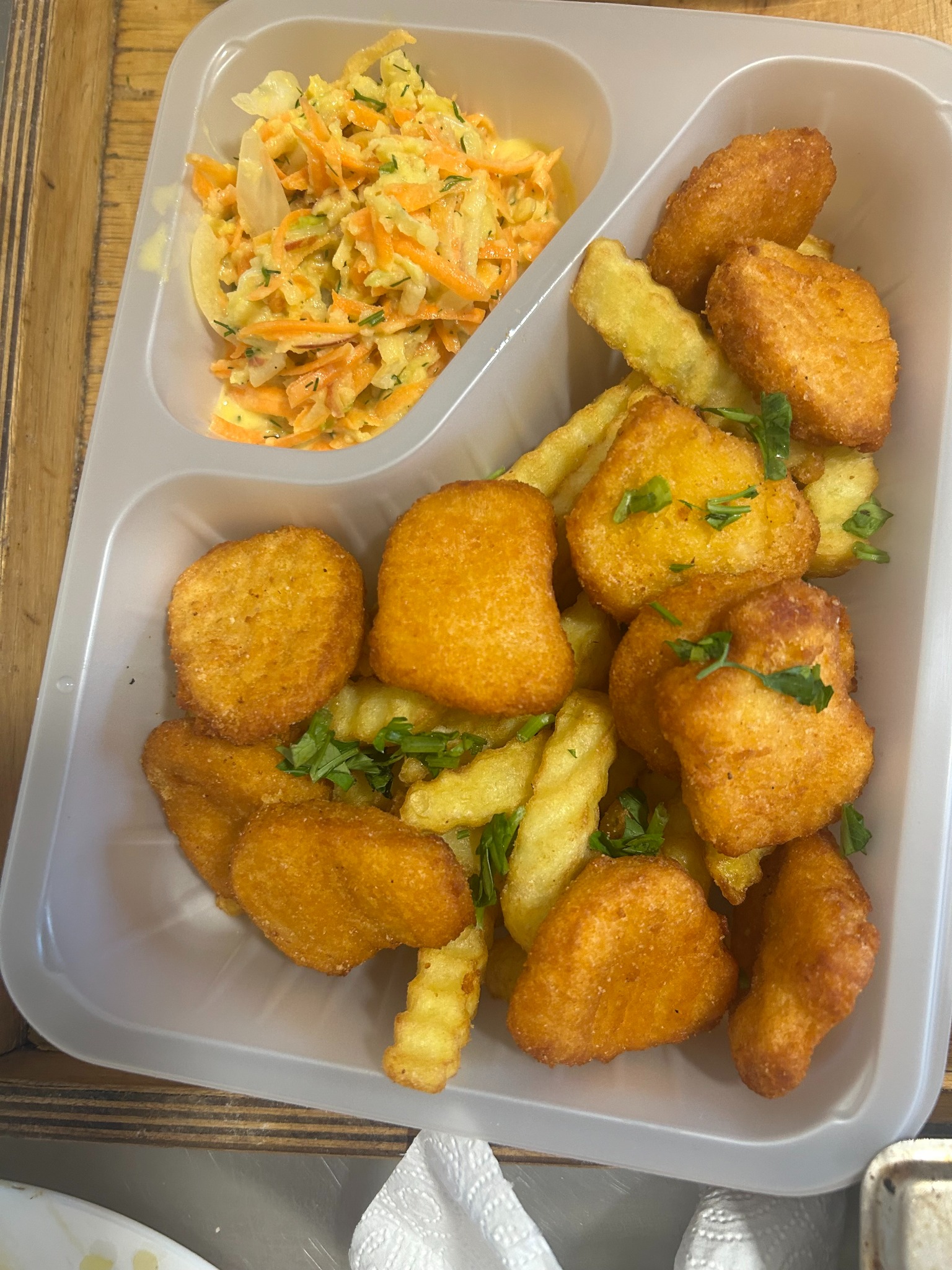 Porcja nuggetów z frytkami i surówką w plastikowym pojemniku cateringowym, posypana zieloną pietruszką, na drewnianym stole. Gotowy zestaw obiadowy.