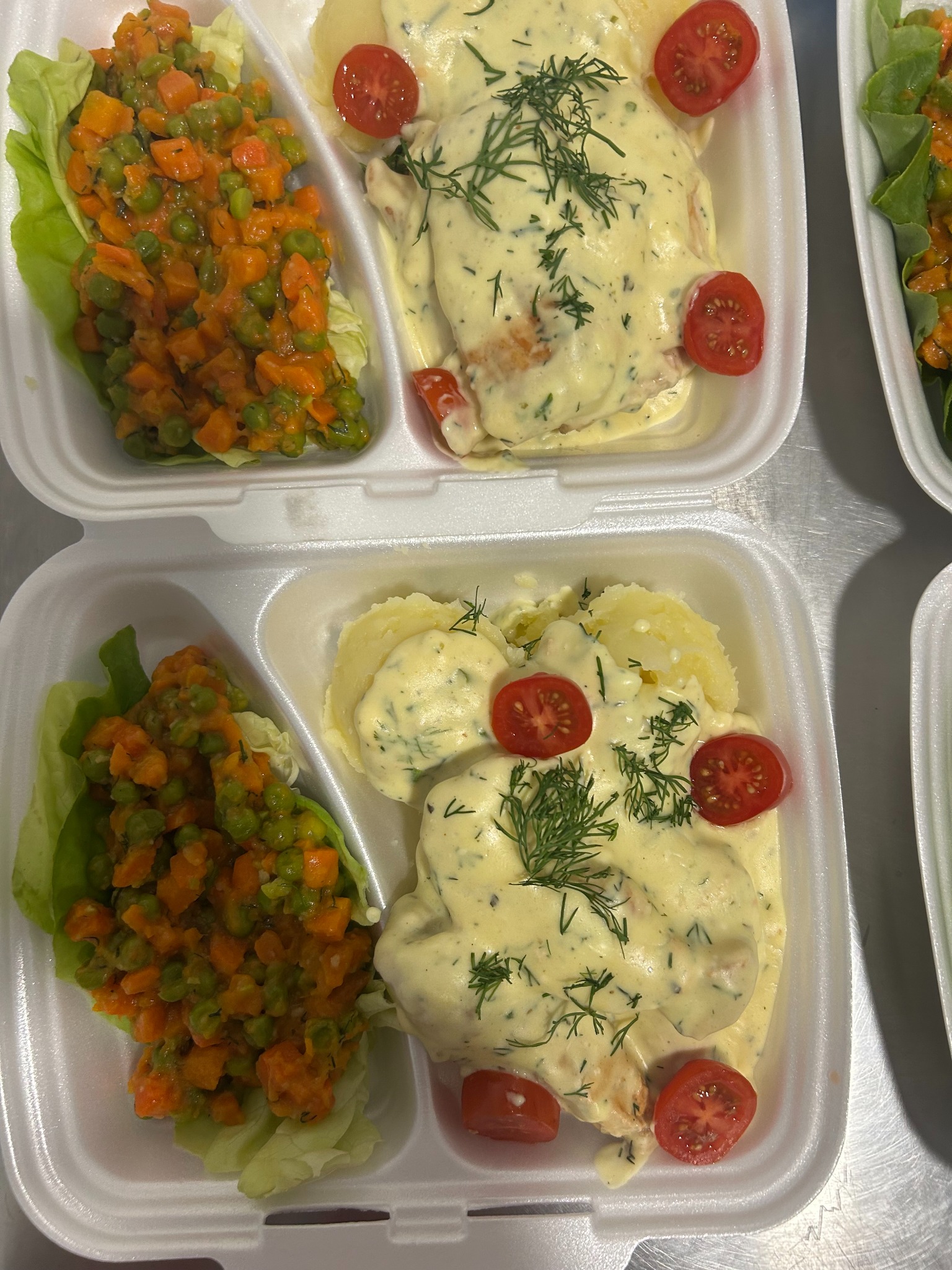 Dania cateringowe w białych pojemnikach: sałatka warzywna na liściach, ryba w sosie koperkowym z ziemniakami, udekorowana pomidorkami cherry i koperkiem.