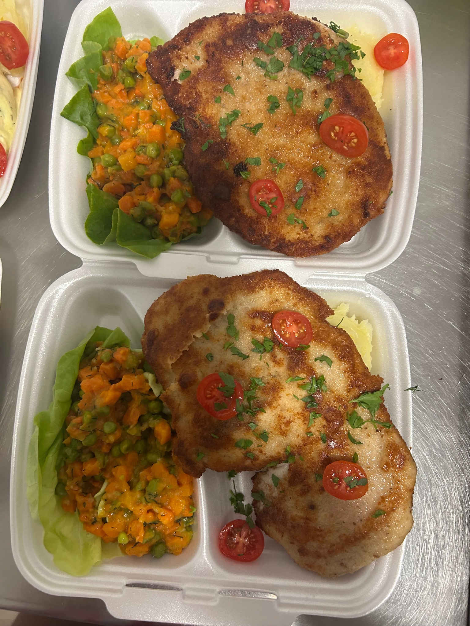 Dwa białe pojemniki z kotletami, sałatką jarzynową i ziemniakami, udekorowane pomidorkami koktajlowymi i natką pietruszki, gotowe do transportu.