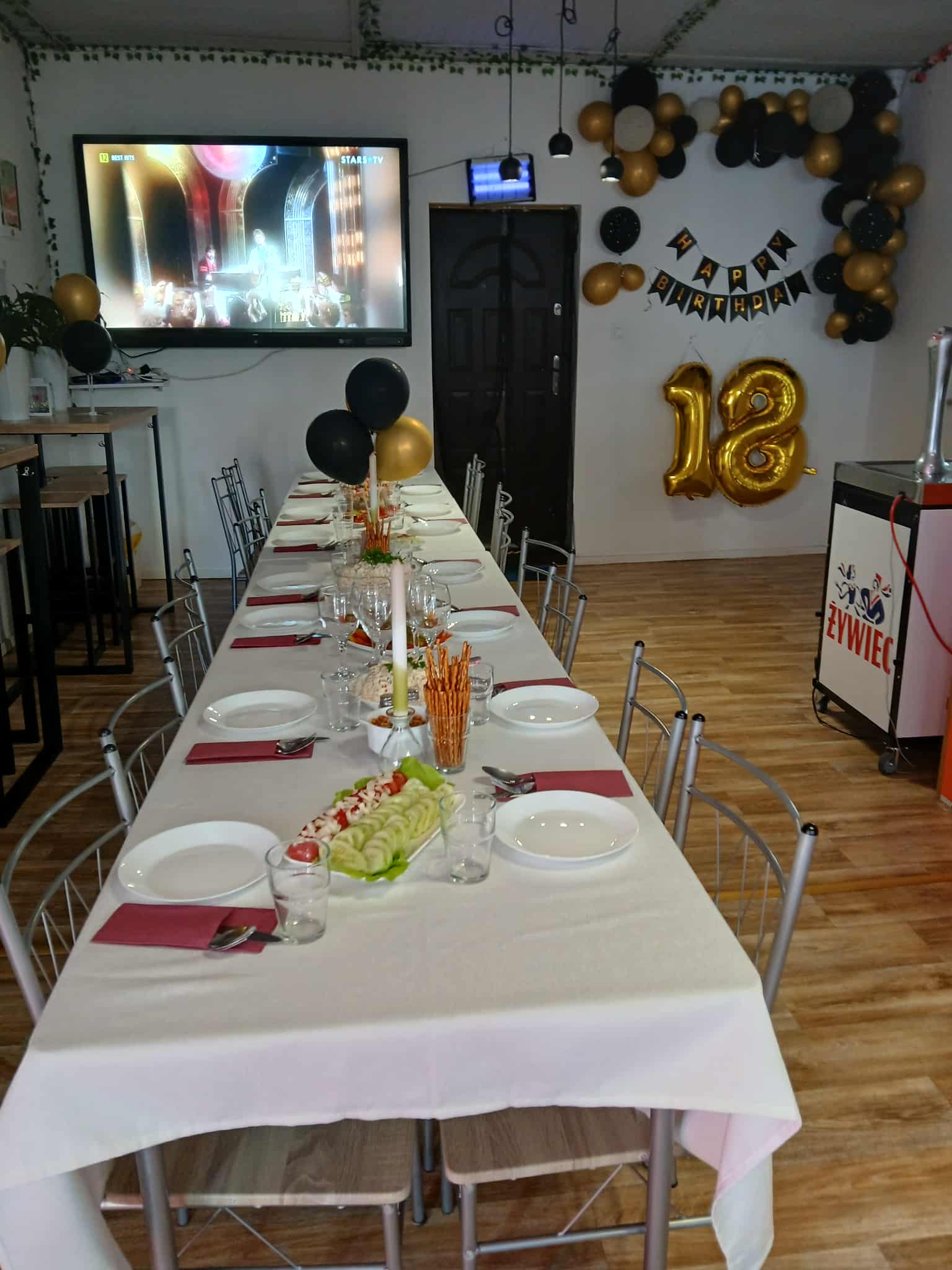 Uroczysty stół na 18 urodziny: biały obrus, zastawa, dekoracje balonowe w złocie i czerni, napis 'Happy Birthday' i cyfra '18'. W tle włączony telewizor.