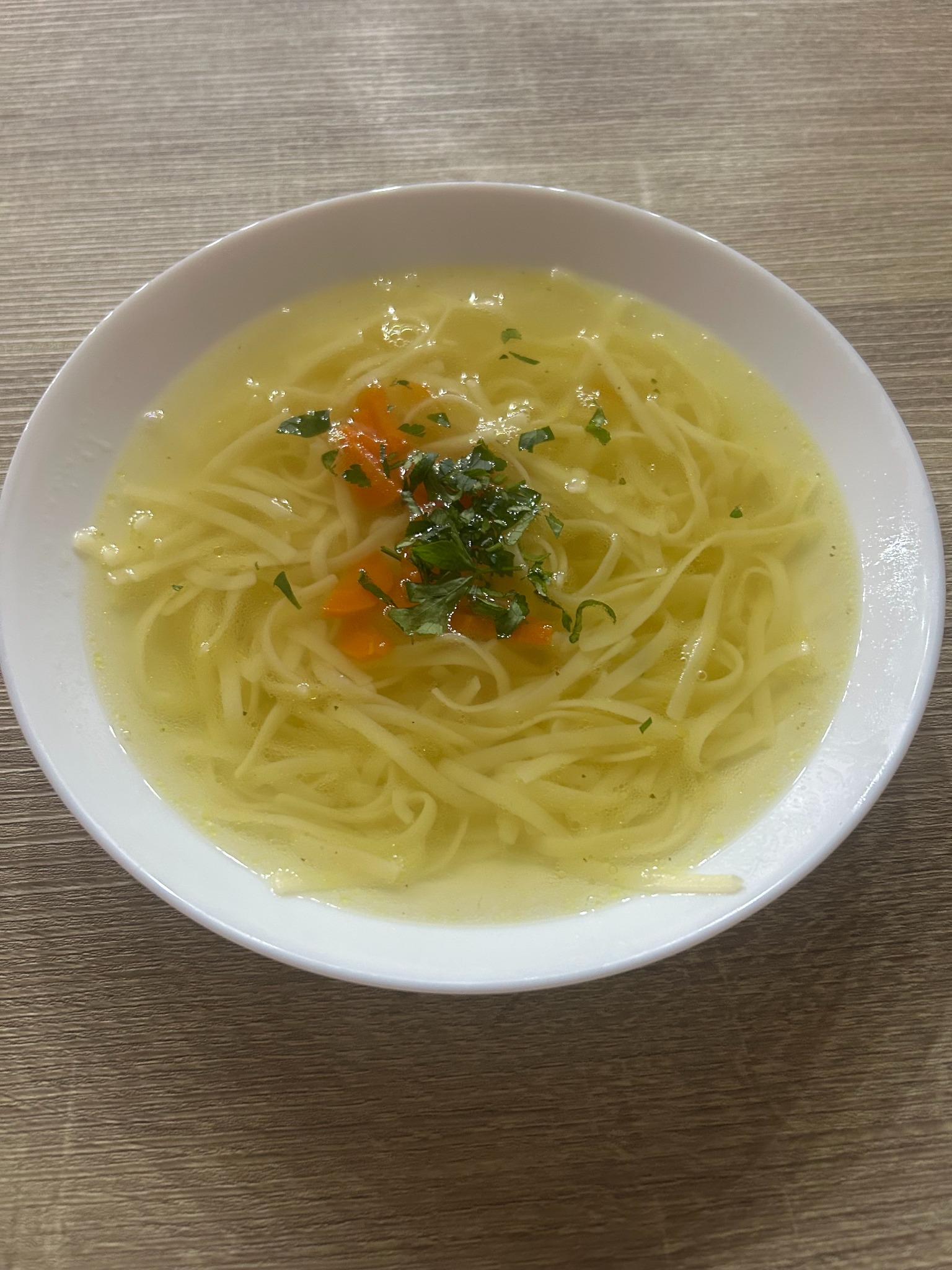 Aromatyczny rosół z makaronem, marchewką i zieloną pietruszką w białym talerzu na drewnianym stole. Idealny na catering firmowy lub świąteczny.