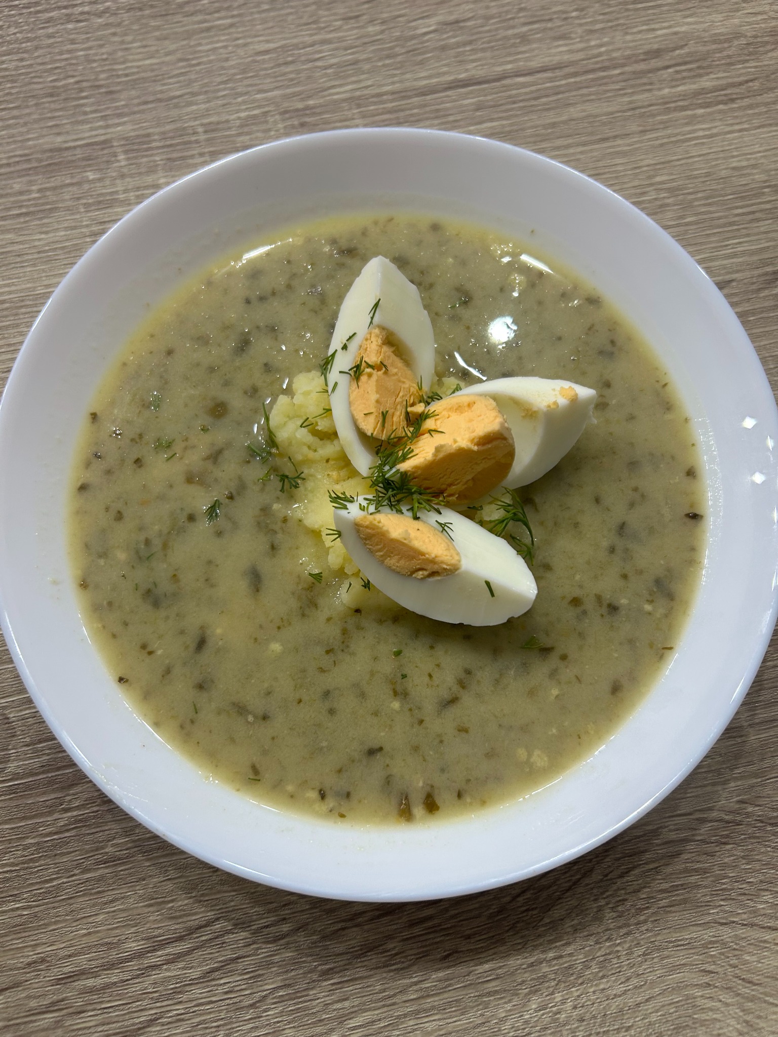 Żurek w białym talerzu, udekorowany ćwiartkami jajka i świeżym koperkiem, na drewnianym stole. Zupa o kremowej konsystencji, gotowa do podania.