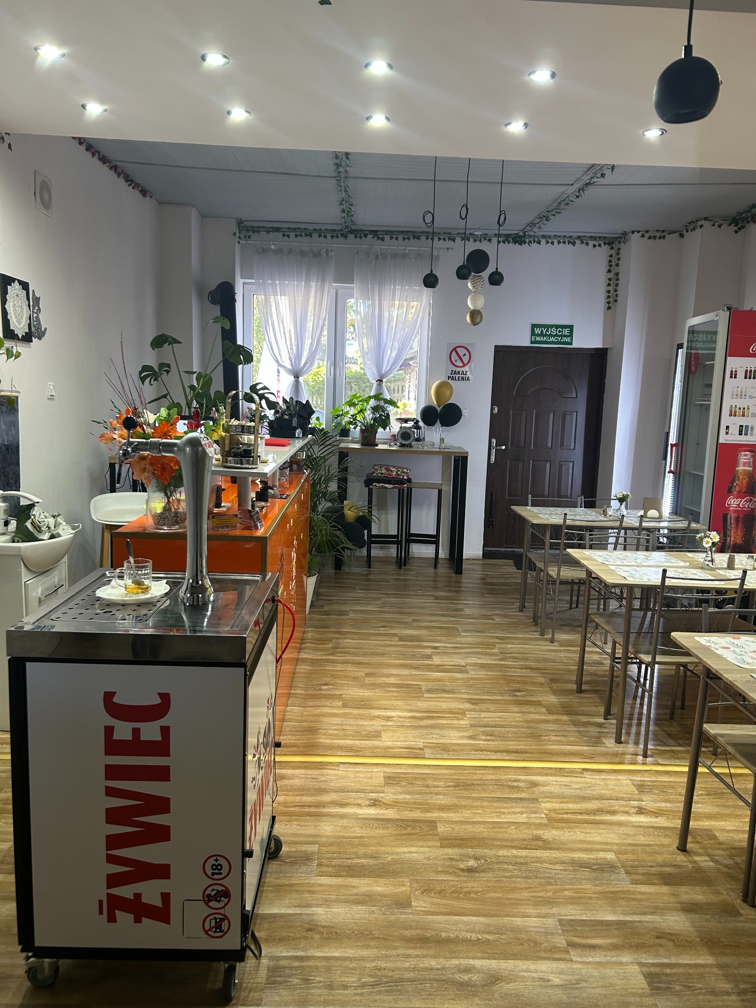 Wnętrze lokalu gastronomicznego z barem, stolikami i dekoracjami roślinnymi. Widoczny dystrybutor piwa Żywiec i lodówka Coca-Cola. Przytulna aranżacja z elementami rustykalnymi.