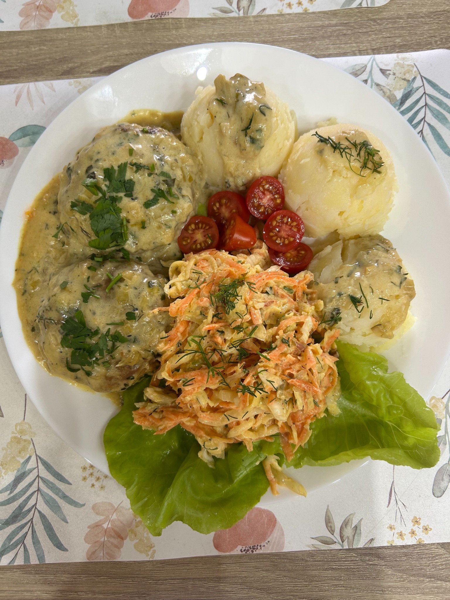 Apetyczny talerz z klopsikami w sosie, ziemniakami, surówką z marchwi i pomidorkami koktajlowymi, udekorowany zieleniną, na tle stołu z motywem roślinnym.