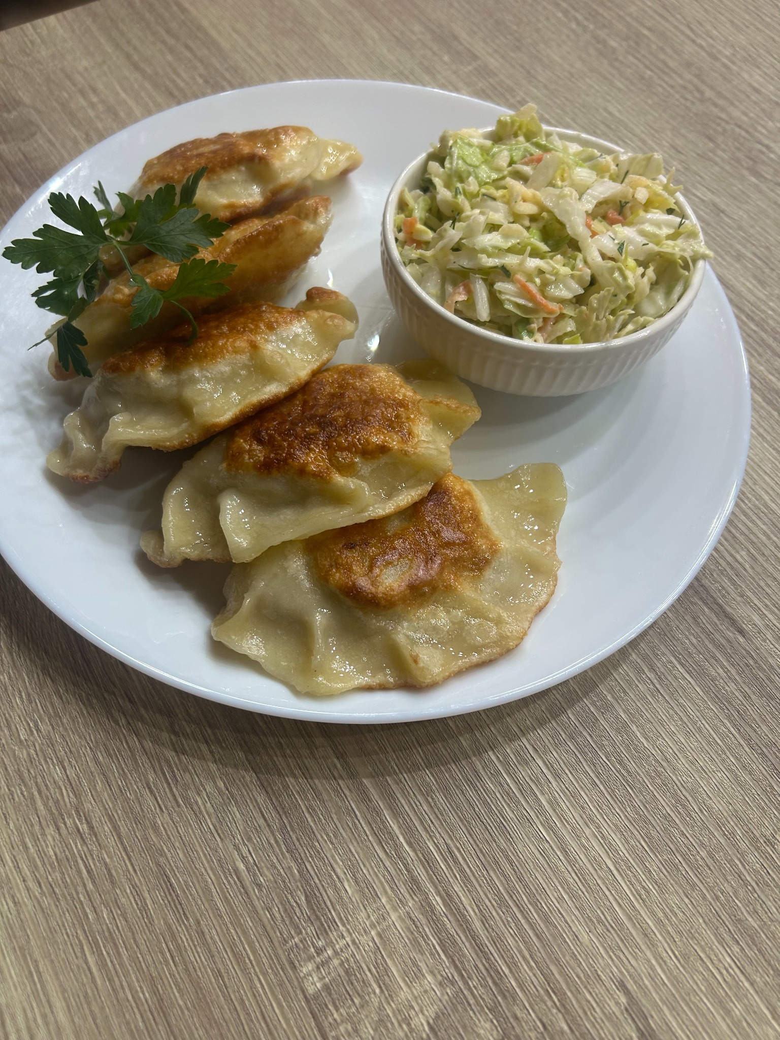 Apetyczne, smażone pierogi z kapustą i grzybami, udekorowane zieloną pietruszką, podane z surówką colesław na białym talerzu, na drewnianym stole.