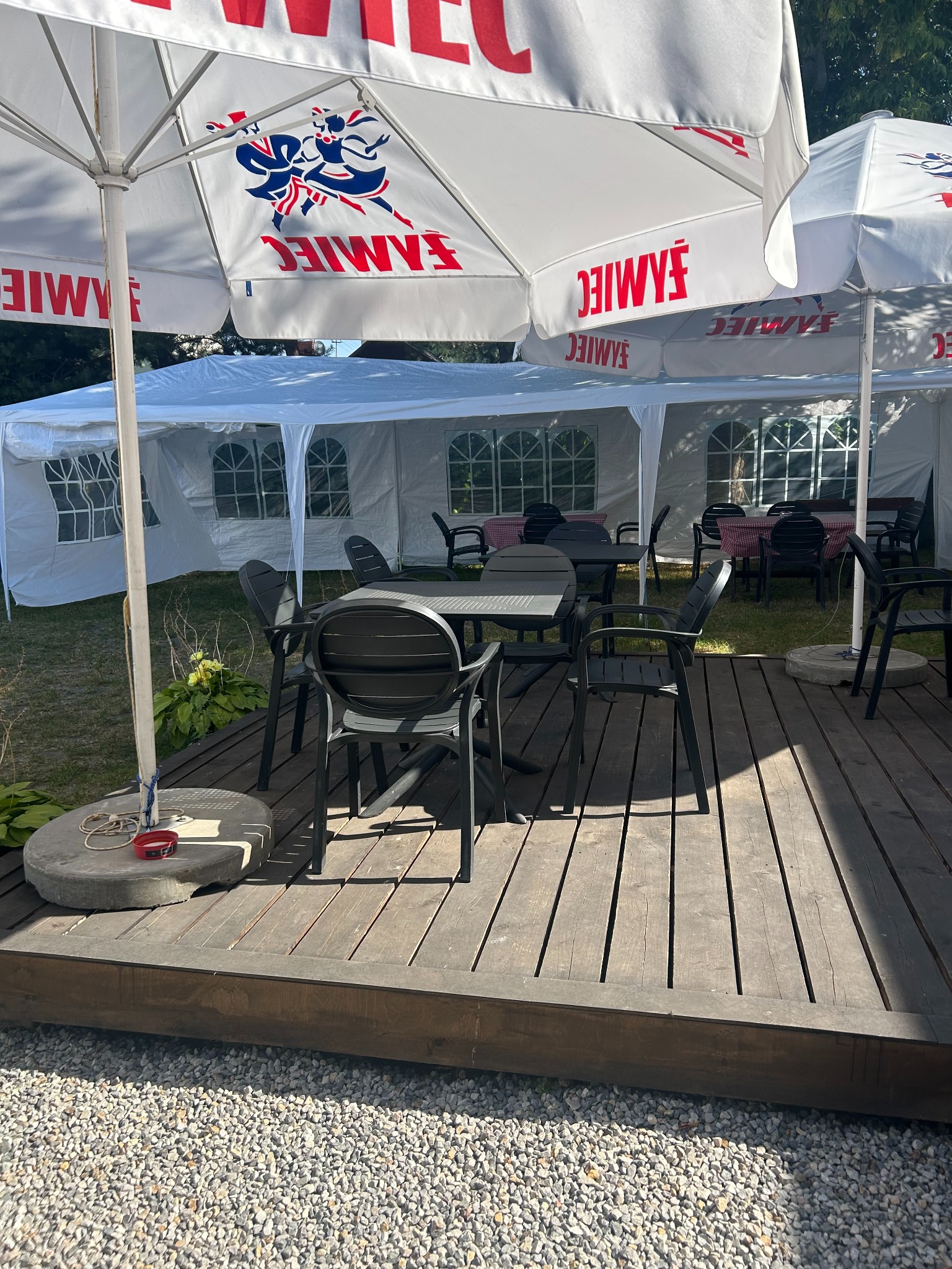 Ustawione stoły i krzesła pod parasolami z logo na drewnianym podeście, w tle biały namiot. Przygotowania do imprezy plenerowej.