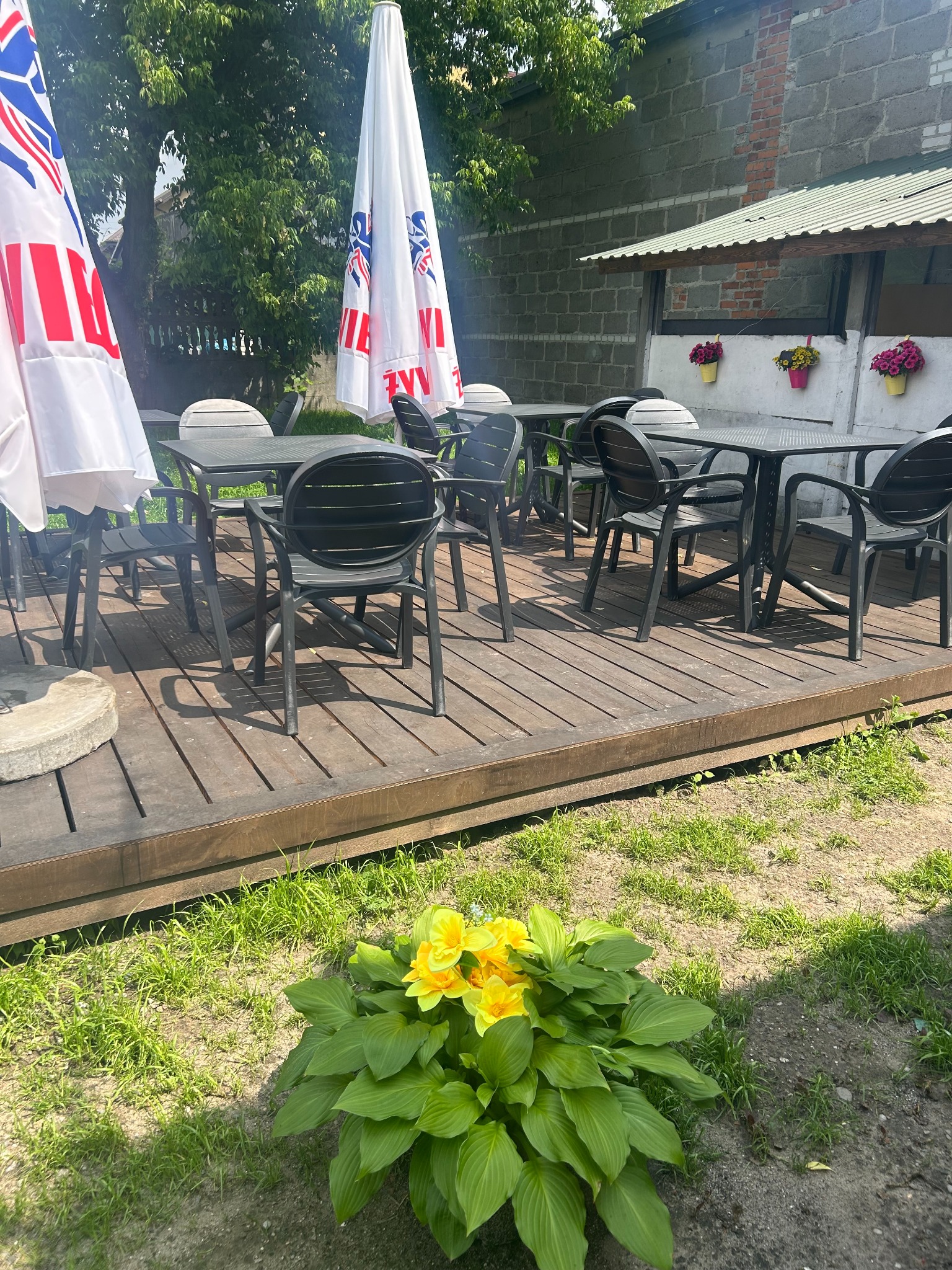 Zewnętrzny ogródek restauracyjny z drewnianym podestem, czarnymi stolikami i krzesłami, parasolami z logo i kwiatami w doniczkach na ścianie budynku.