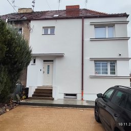 Nowy start - Odnowiony, dwukondygnacyjny dom z białymi ścianami i schodami wejściowymi, widoczny z zewnątrz, z czarnym samochodem na pierwszym planie i zielenią w tle.