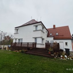 Nowy start - Biały dom z czerwoną dachówką i drewnianym tarasem. Widok z ogrodu, pochmurne niebo. Elementy dekoracyjne na tarasie i w ogrodzie. Nowe wykończenie elewacji.