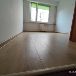 Nowy start - Jasne panele podłogowe w pokoju po remoncie w Nysie. Widok na okno z zieloną roletą i grzejnik. Świeżo wykończone wnętrze z listwami przypodłogowymi.