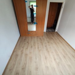 Nowy start - Jasna podłoga z paneli, drewniana szafa z lustrem zabezpieczona taśmą, białe ściany i widok na sąsiednie pomieszczenie. Ujęcie szerokokątne.
