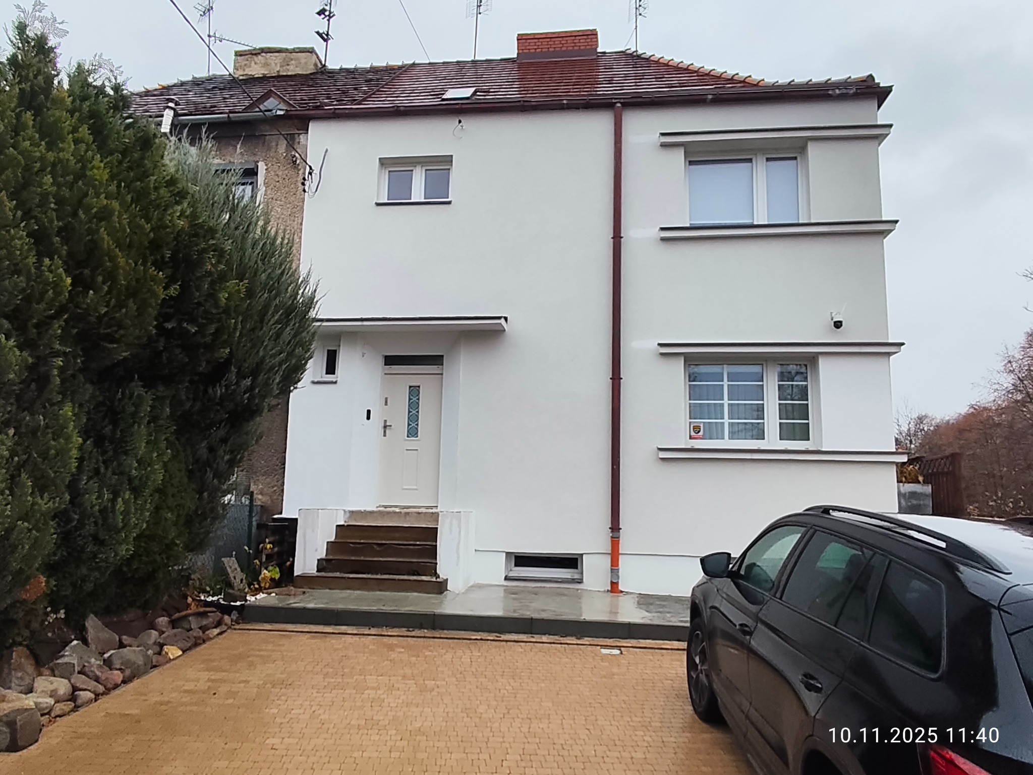 Odnowiony, dwukondygnacyjny dom z białymi ścianami i schodami wejściowymi, widoczny z zewnątrz, z czarnym samochodem na pierwszym planie i zielenią w tle.