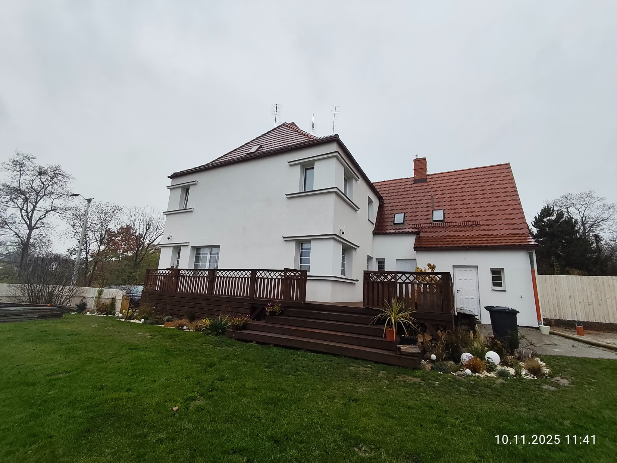Biały dom z czerwoną dachówką i drewnianym tarasem. Widok z ogrodu, pochmurne niebo. Elementy dekoracyjne na tarasie i w ogrodzie. Nowe wykończenie elewacji.